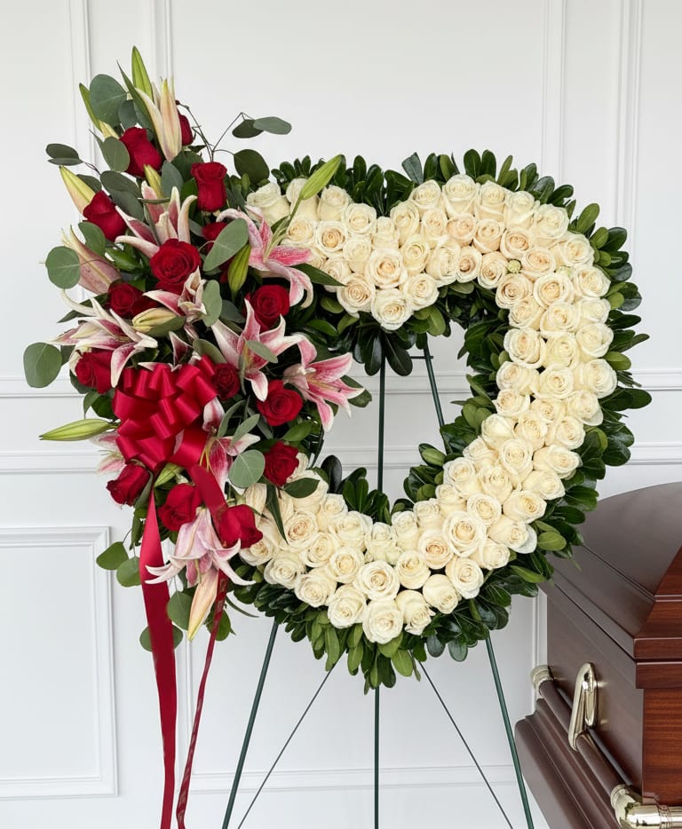 Eternal Devotion Heart - A stunning expression of everlasting love, this luxurious heart-shaped wreath is crafted with an abundant display of soft white roses, symbolizing purity, honor, and eternal remembrance. Accented with a dramatic cluster of rich red roses and elegant lilies, this design beautifully represents deep love, respect, and devotion.  Lush greenery and flowing ribbon details add depth and movement, creating a striking and sophisticated tribute. The heart shape serves as a powerful symbol of an unbreakable bond, making this piece a meaningful way to honor a life that will always be remembered.  Available in multiple sizes, this arrangement offers a refined and heartfelt expression of sympathy, love, and enduring connection.