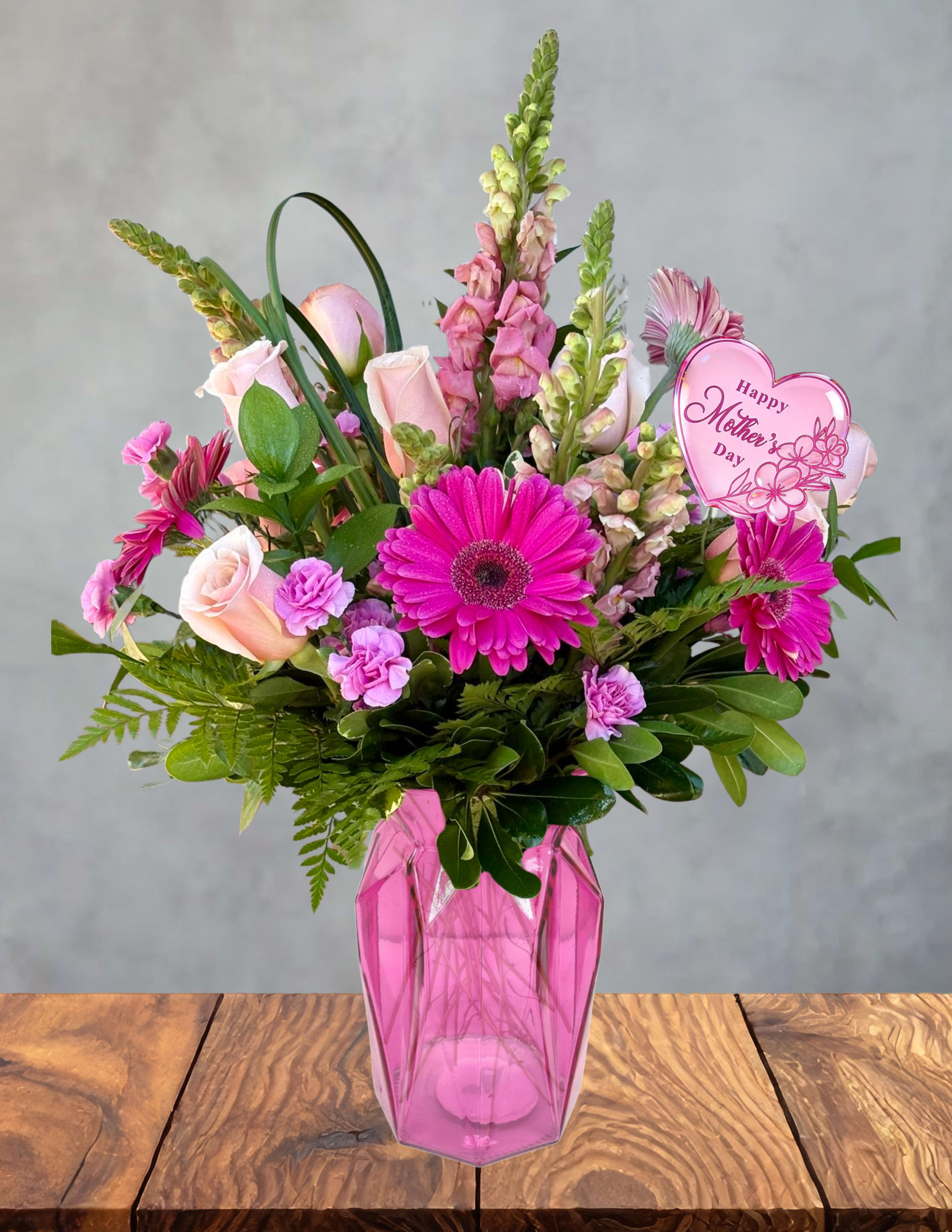 Pink Meadow Mama - A lively burst of color made to celebrate the mama who brings joy wherever she goes.  Pink Meadow Mama shines with vibrant pink roses, bold magenta gerberas, soft snapdragons, and playful purple blooms, creating a cheerful mix that feels fresh, modern, and full of heart. The lush greens add movement and texture, giving the bouquet that “wild meadow” energy while still feeling polished and intentional.  A sweet pink heart pick adds a festive Mother’s Day touch, making the design feel personal and full of affection. Presented in a chic pink geometric vase, this arrangement radiates confidence, warmth, and the bright, uplifting spirit of a mama who deserves to be celebrated in full color.  A perfect choice for the mom who loves bold beauty, joyful moments, and flowers that make a statement.