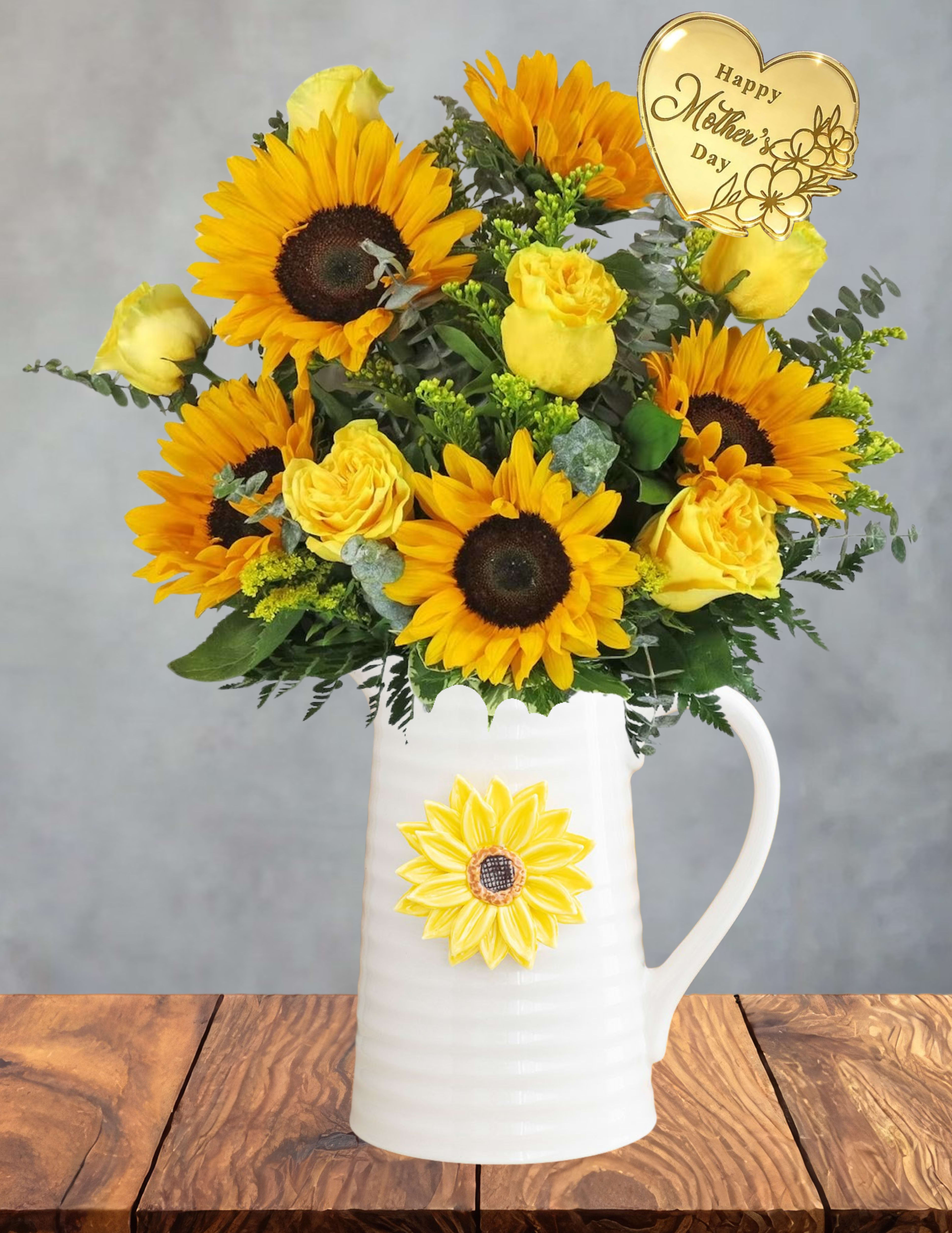 Sunshine for Mama - A cheerful burst of golden blooms made to brighten her day the way she brightens yours.  Sunshine for Mama glows with radiant sunflowers, soft yellow roses, and fresh greenery, creating a warm, uplifting mix that feels like a beam of morning light. The vibrant yellows bring instant joy, while the natural textures give the arrangement that breezy, meadow‑fresh charm.  A gold heart pick adds a sweet Mother’s Day touch, making the design feel personal, celebratory, and full of love. Presented in a charming white pitcher adorned with a sunflower motif, this keepsake vessel extends the gift long after the flowers have faded — perfect for future blooms or as a cozy home accent.  Bright, heartfelt, and full of gratitude, this arrangement is a beautiful way to say “Mama, you are my sunshine.”
