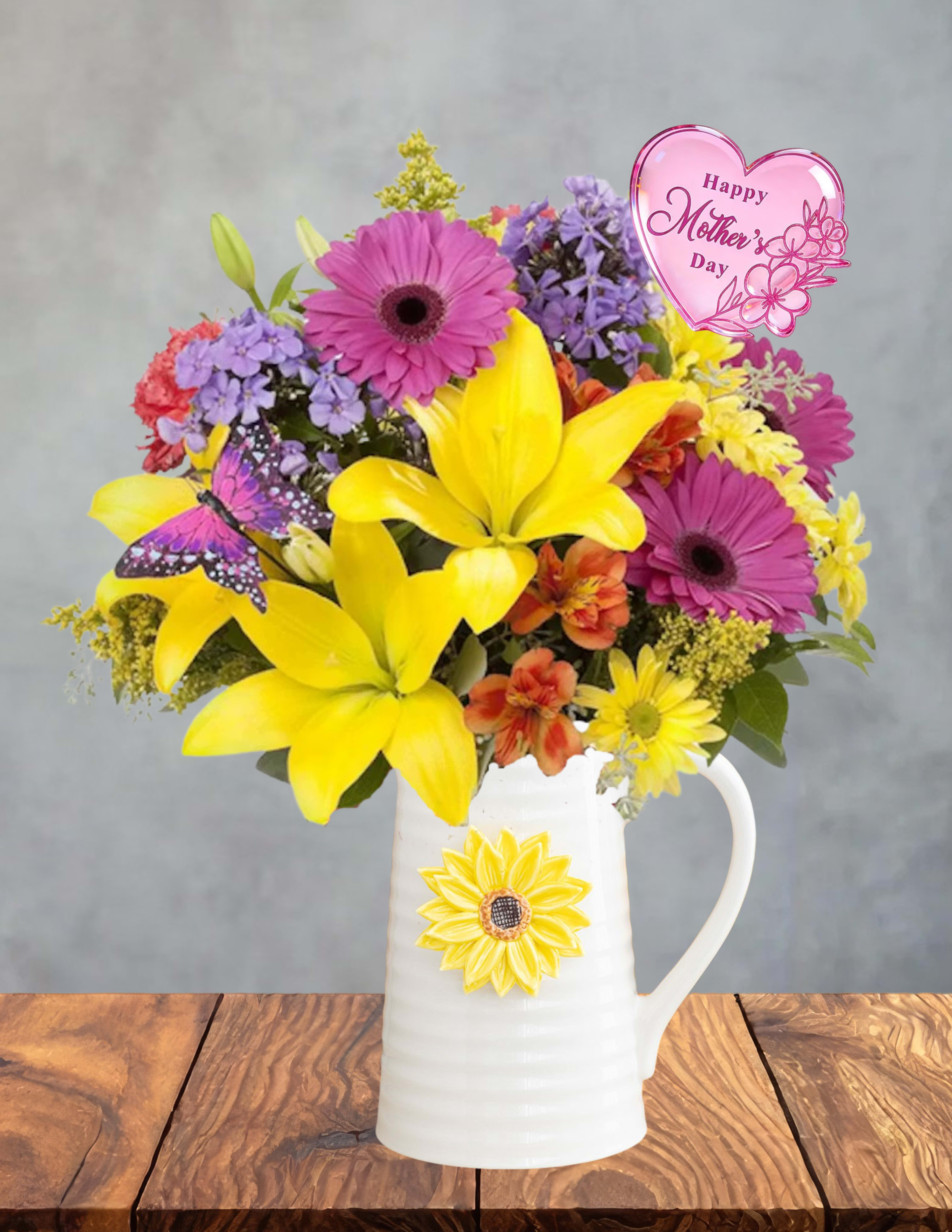 Mom’s Joyful Pitcher - A colorful celebration of the mom who fills every day with joy.  Mom’s Joyful Pitcher bursts with vibrant yellow lilies, playful purple daisies, bold pink gerberas, and touches of orange and red blooms, creating a lively, rainbow‑bright mix that feels festive, warm, and full of love. Each flower adds its own spark of happiness, coming together like a little garden party made just for her.  A sweet Mother’s Day heart accent adds a celebratory touch, while the white ceramic pitcher — adorned with a sunny sunflower — becomes a charming keepsake she can enjoy long after the flowers have faded. Whether she uses it for fresh blooms, kitchen décor, or simply as a reminder of your love, it’s a gift that keeps giving.  Joyful, colorful, and overflowing with appreciation, this arrangement is the perfect way to say “Mom, you brighten my world.”