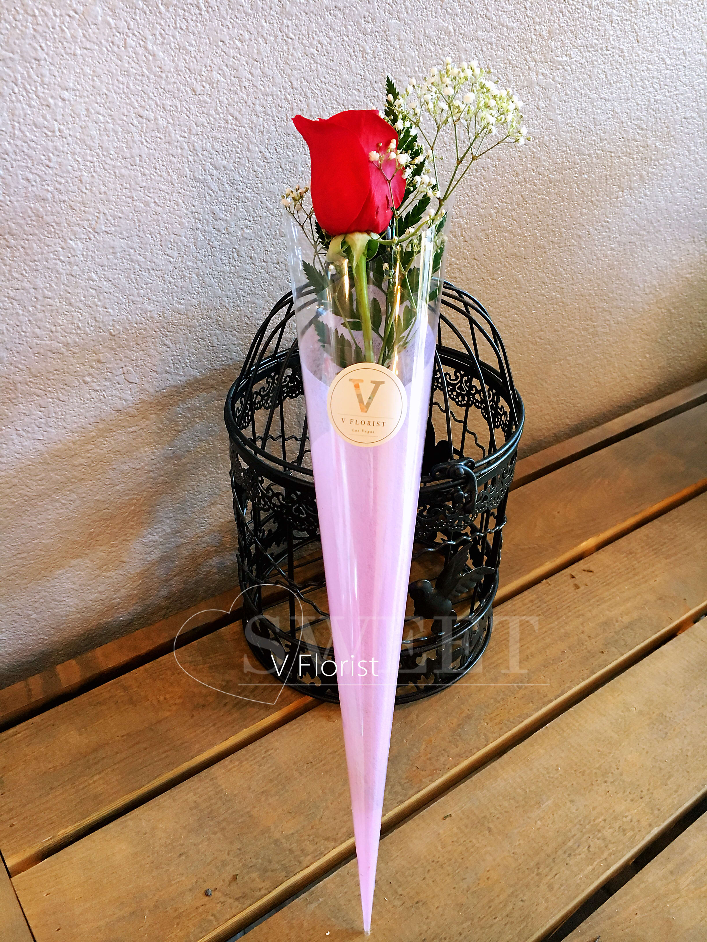 Single red rose with white filler flowers wrapped in a pink cone sleeve.