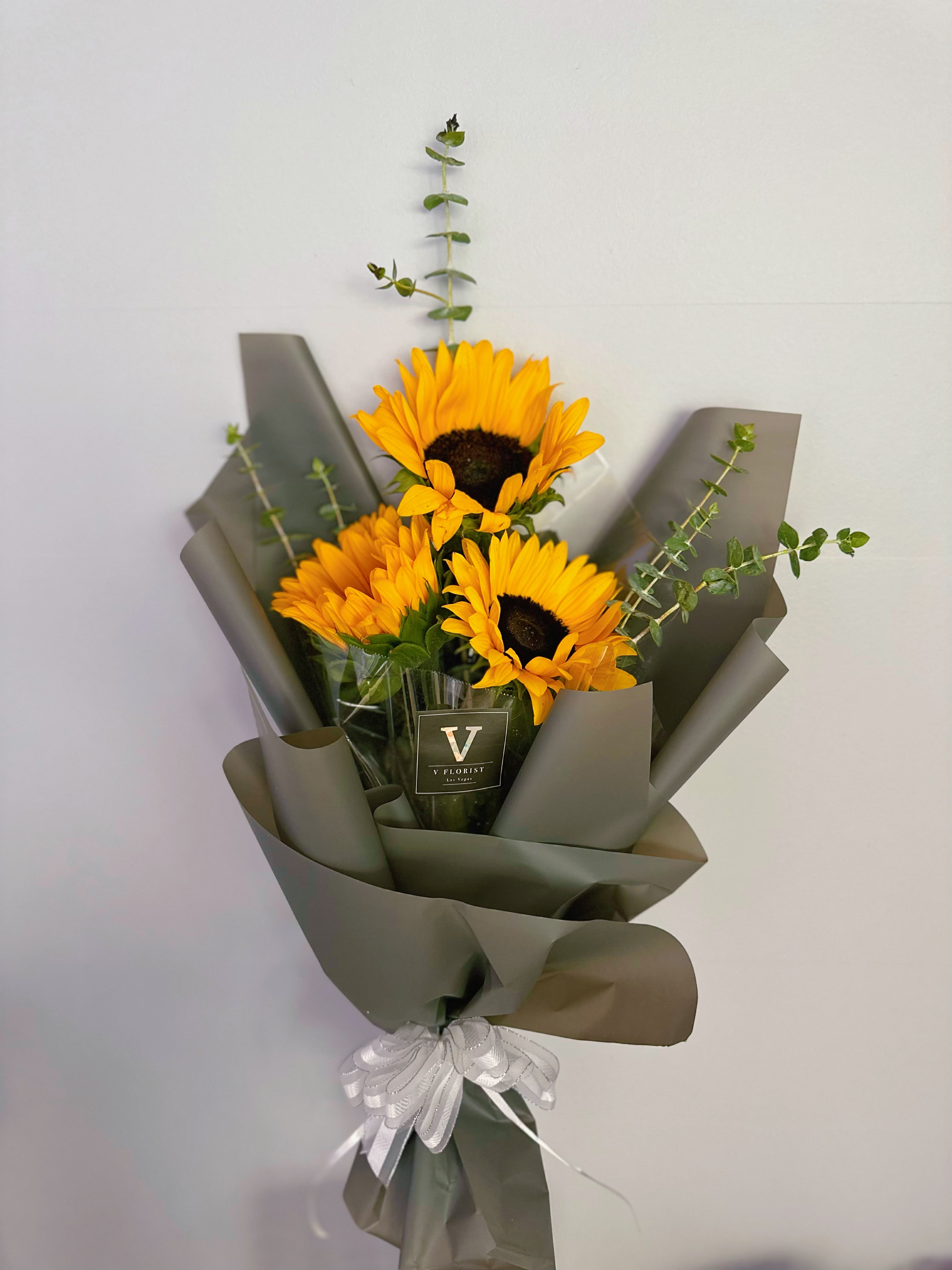 Bouquet of yellow sunflowers with tall eucalyptus stems wrapped in dark paper with a white ribbon bow