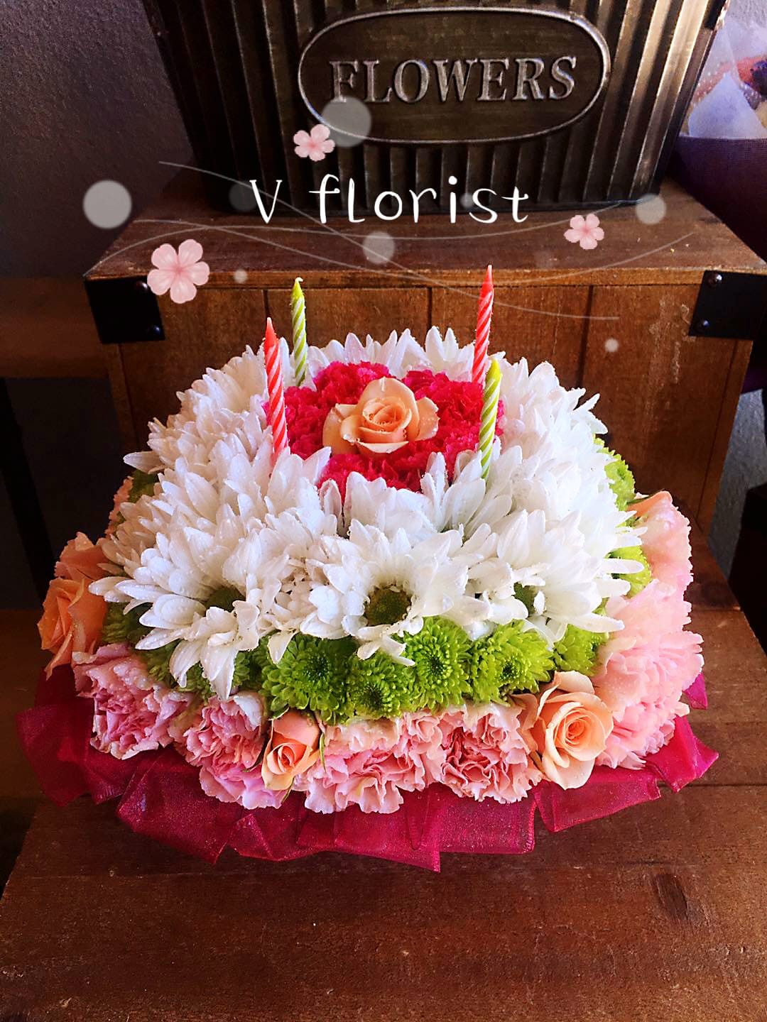 Round floral cake of white daisies, pink carnations, roses, and green mums topped with striped candles.