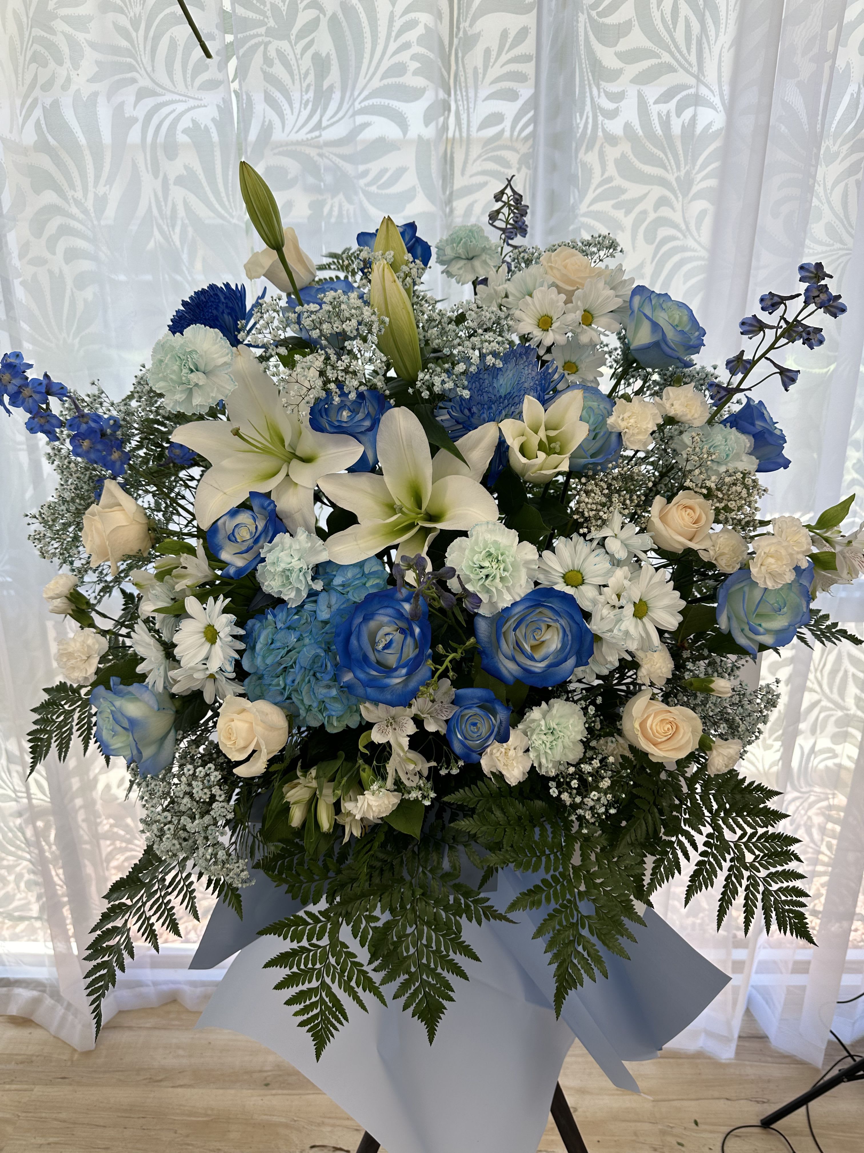 Tall blue and white standing flower arrangement with lilies, roses, and daisies on a tripod stand