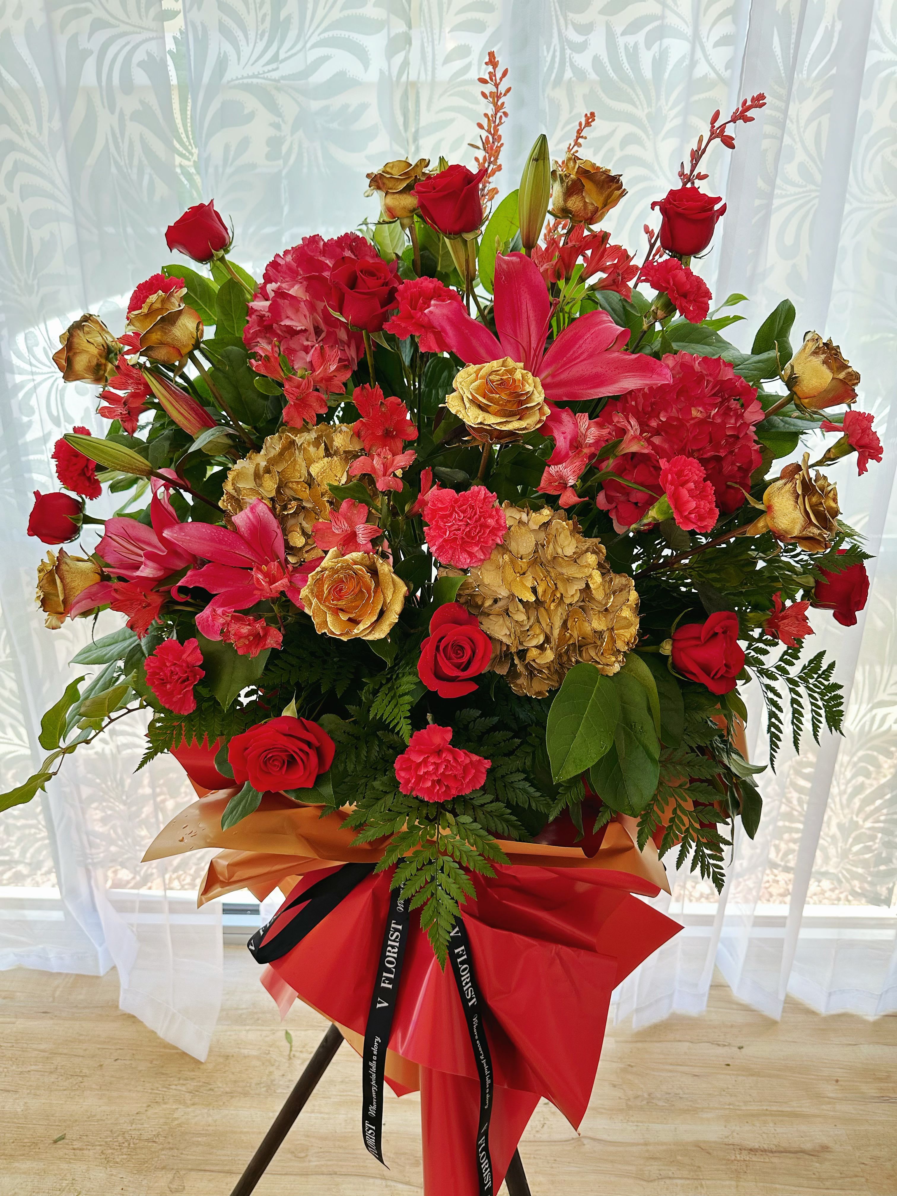 Tall standing arrangement of red roses, lilies, carnations and gold-toned blooms wrapped in red and gold paper.