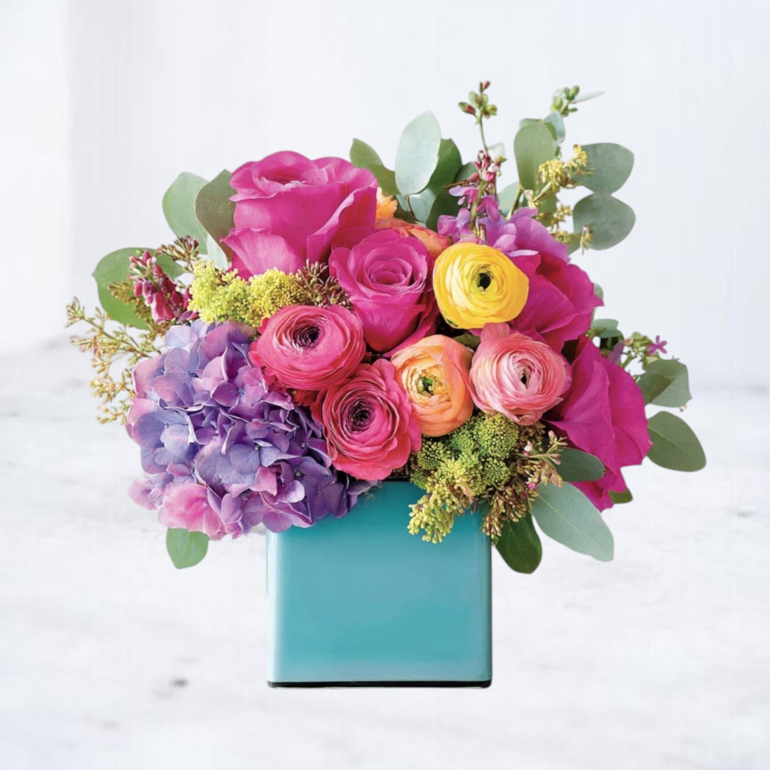 Picturesque - Vivid pink roses and ranunculus take center stage in this birthday flower arrangement, surrounded by peach and yellow ranunculus for warm contrast-perfect for a birthday bouquet or a bright table accent. A cloud-like head of purple hydrangea anchors one side while blue and orange notes lift the palette; the overall effect reads joyful and modern, made to make the person you're thinking of feel celebrated and truly seen.  This arrangement combines hydrangeas, ranunculus, and roses in shades of pink, purple, yellow, orange, and blue, accented with small green filler flowers and rounded foliage for a lush, layered look. Blooms are arranged compactly in a smooth turquoise ceramic cube vase that adds a playful, contemporary touch and keeps the design feeling fresh on mantels, desks, or dining tables.  Ideal for birthdays, congratulations, special gatherings, or as a cheerful everyday gift, Picturesque arrives ready to enjoy; the compact shape works well for same-day delivery and for brightening small spaces. Gentle daily refreshing of the water and trimming stems every few days will help extend the life of the bouquet.