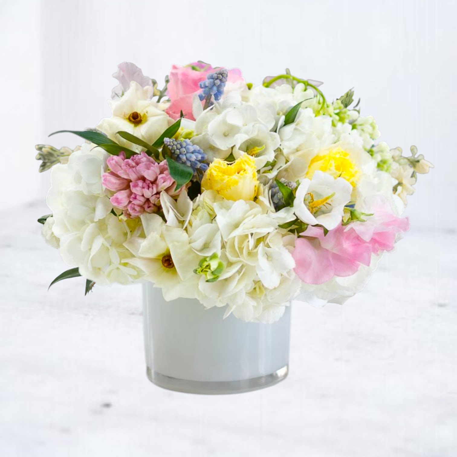 Low pastel arrangement of white hydrangeas with pink, yellow, and blue blooms in a white glass cylinder vase