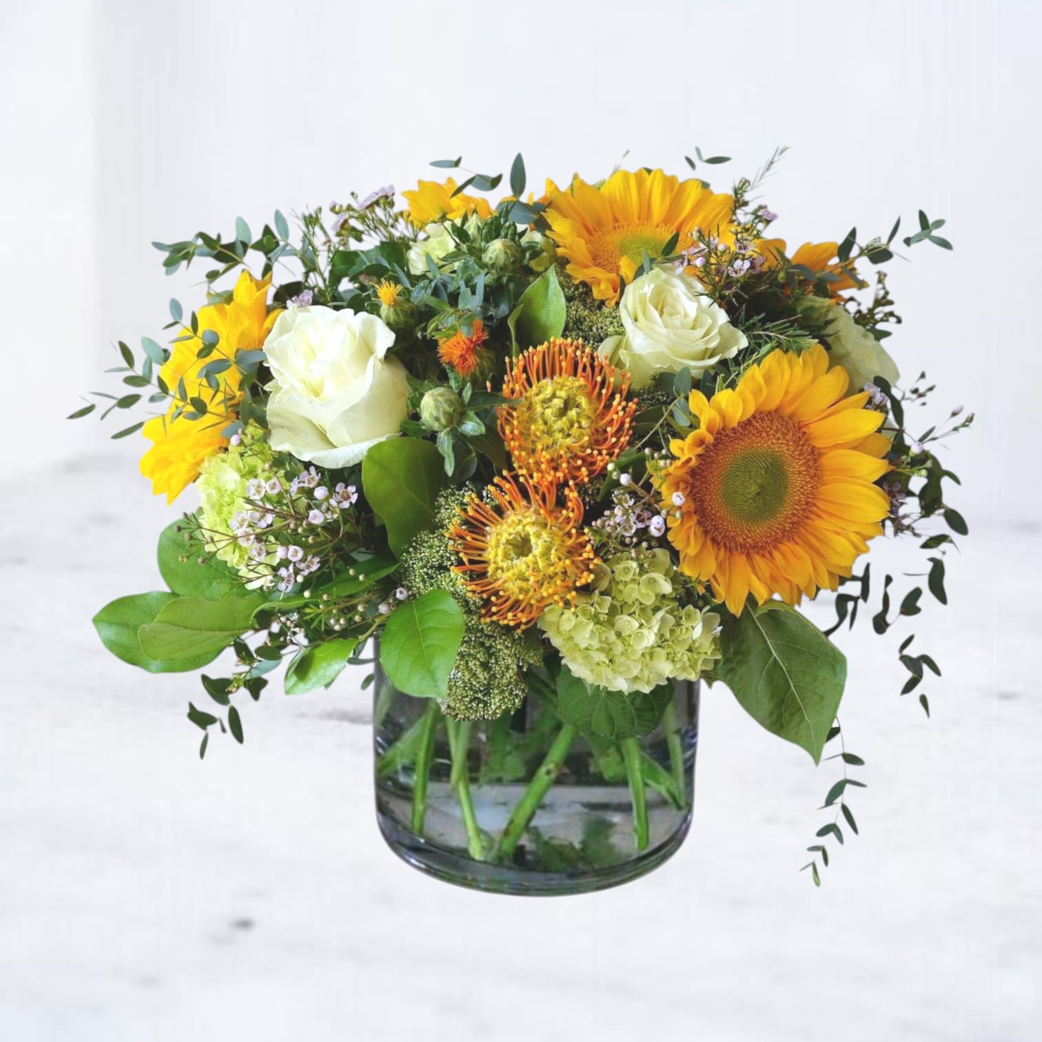 Alderfield - Alderfield is a warm, garden-inspired arrangement featuring vibrant sunflowers, striking pincushion protea, creamy white roses, and seasonal blooms gathered among lush greenery. The golden tones and natural textures create a bright, cheerful design with a relaxed meadow-like feel.  Designed in a clear glass vase with airy movement and rich seasonal color, Alderfield is perfect for birthdays, celebrations, or sending a joyful floral surprise.  Whiskey Wallflower is a design-forward florist serving Magnolia, The Woodlands, Montgomery, Willis, Conroe, Spring, and surrounding Houston-area communities. We specialize in thoughtfully composed, modern floral arrangements with a focus on premium blooms, intentional design, and reliable same-day delivery. Every piece is created with care to make life’s moments feel elevated, personal, and unforgettable.