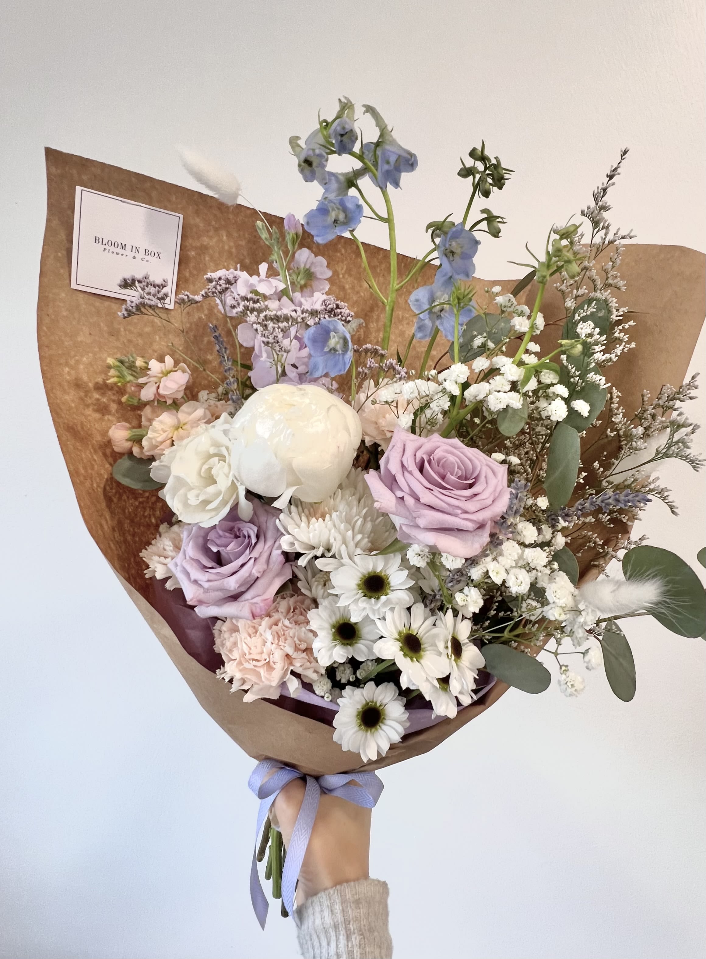 Pastel bouquet of lavender roses, white daisies, and mixed blooms wrapped in brown paper with a purple ribbon.