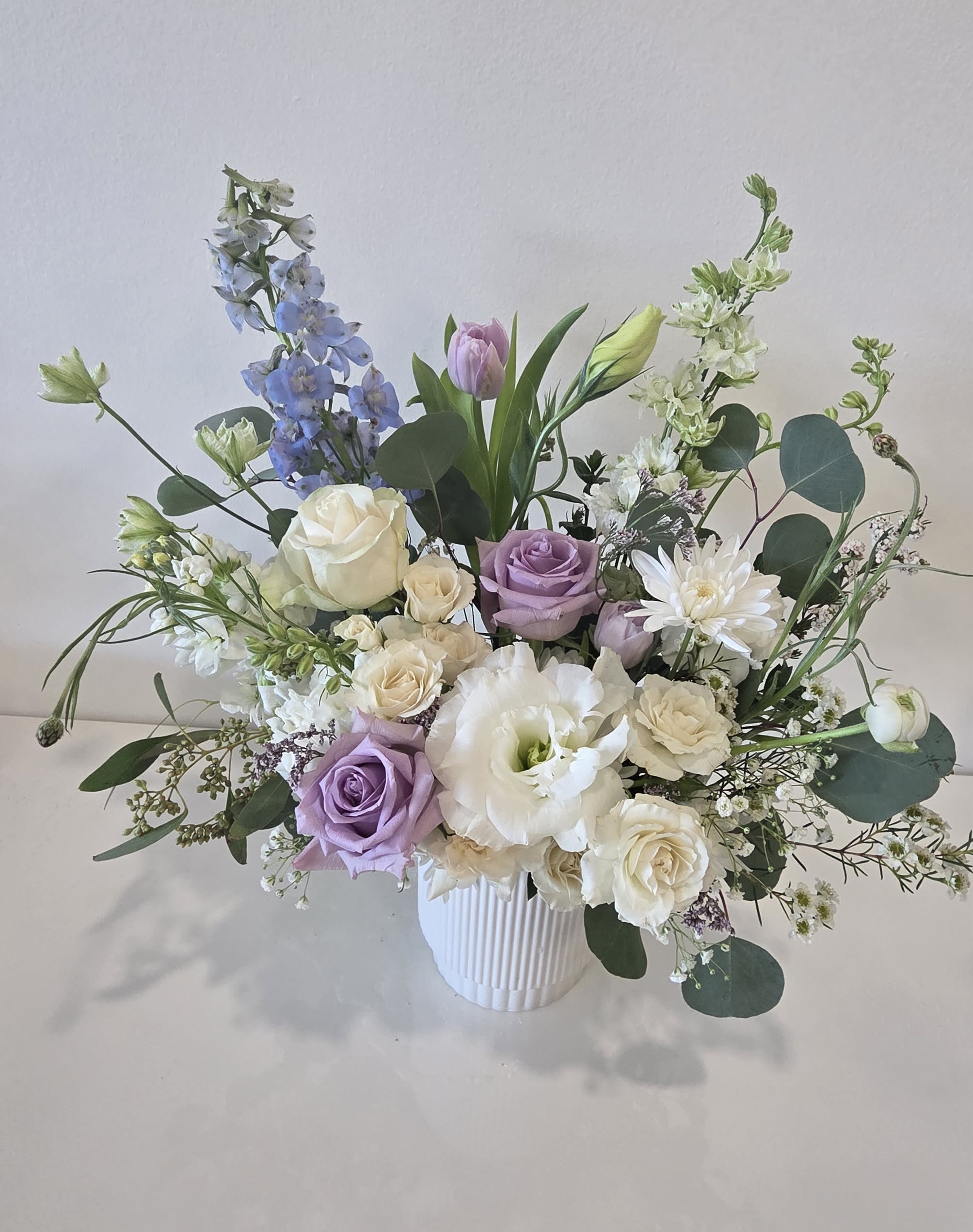 Pastel arrangement of white and lavender roses, lisianthus and delphinium in a white ribbed vase.