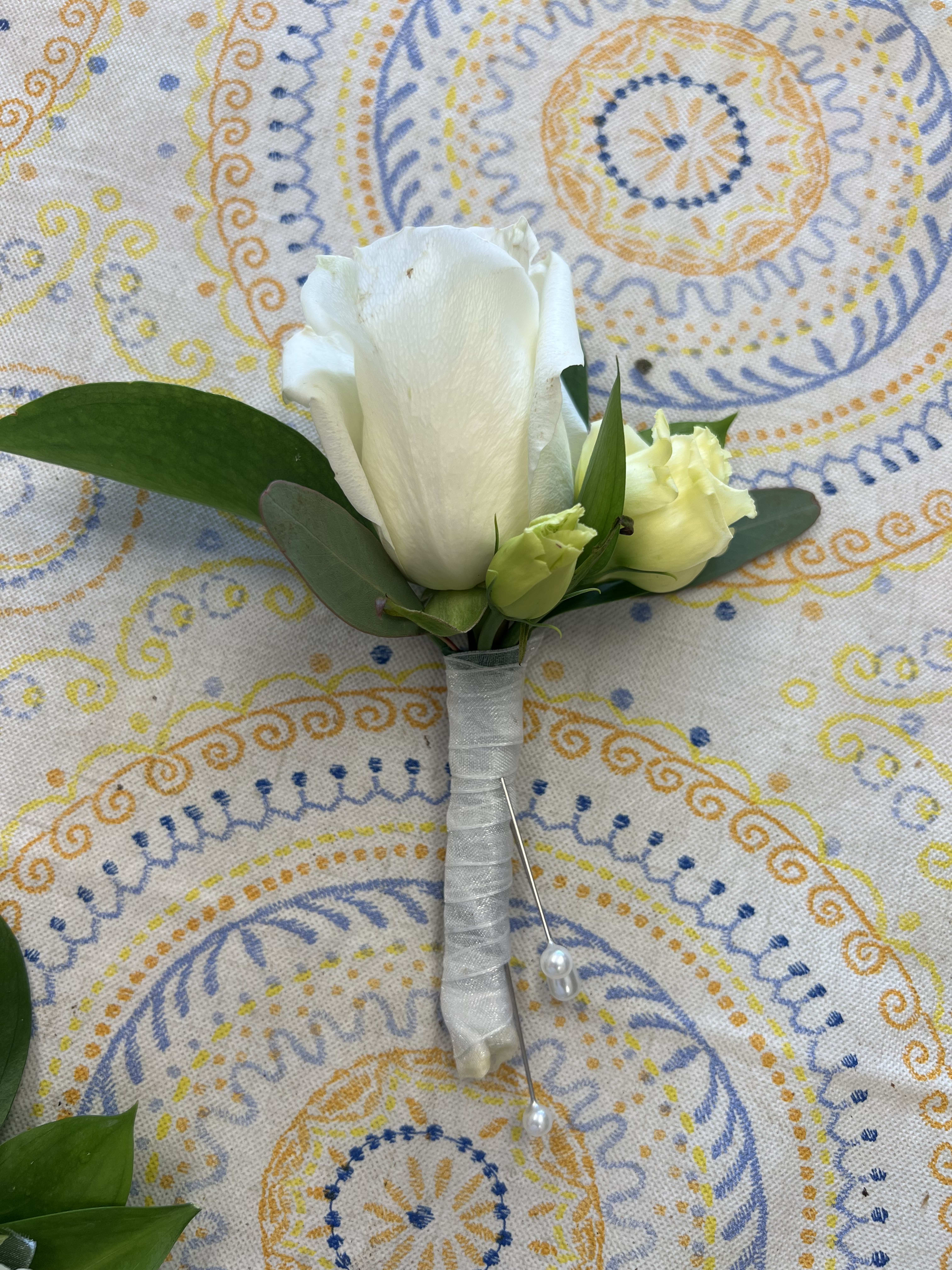 White rose boutonniere with small buds and greenery wrapped in white ribbon with pearl pins on patterned cloth
