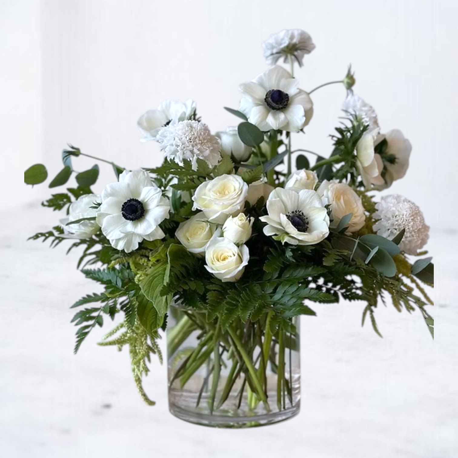 White anemones and roses arranged in a clear glass vase with lush greenery