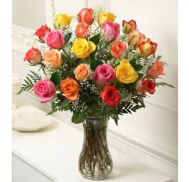 Tall glass vase filled with assorted pink, yellow, orange, and red roses with white filler flowers.