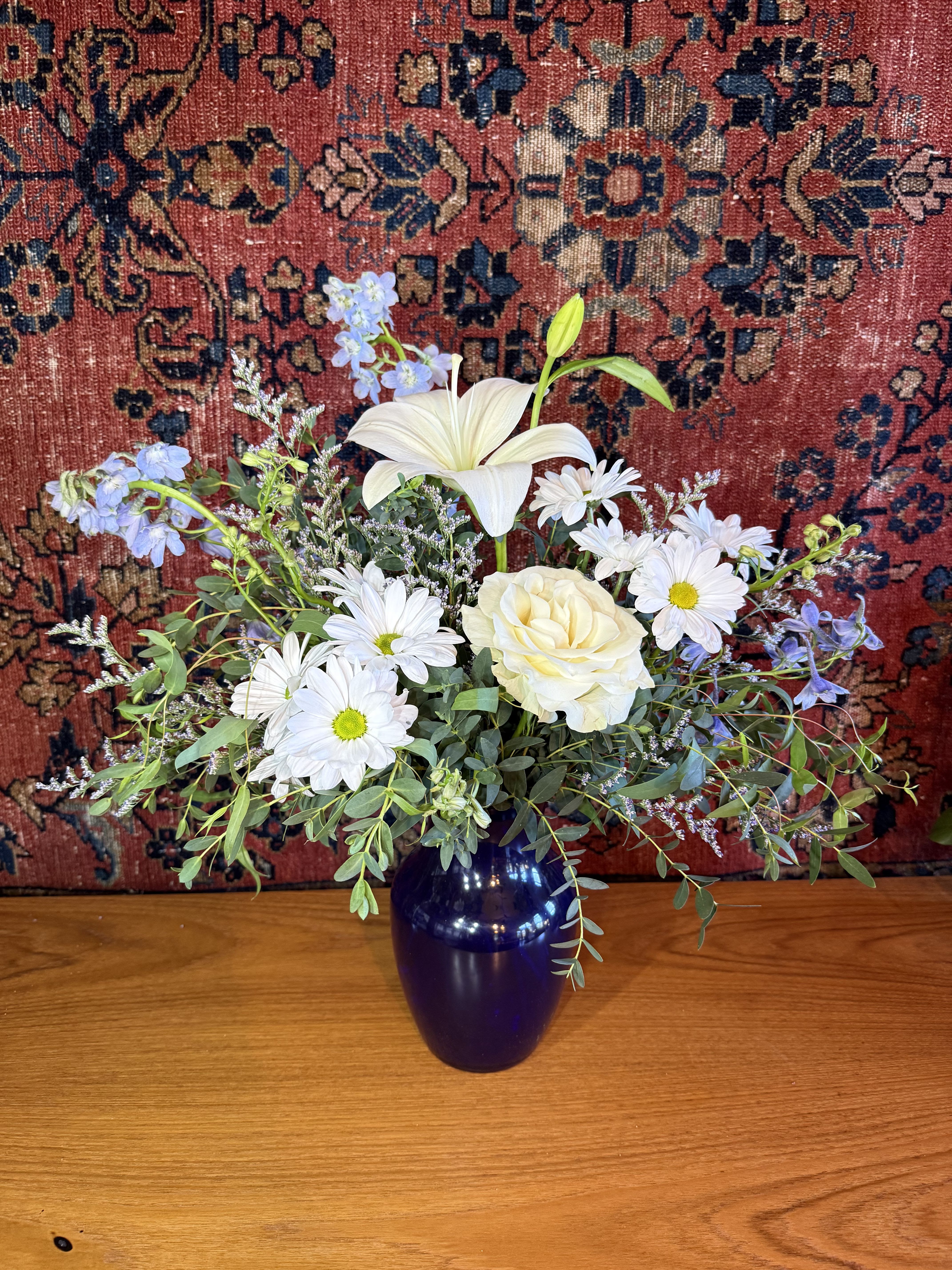 Bella Blue - White lilies take center stage in this calm, elegant flower arrangement-perfect as a sympathy arrangement, a get well bouquet, or a thoughtful Mother's Day gift that communicates quiet support. The rounded mix of white roses, daisy-style blooms, and cushion chrysanthemums is lifted by clusters of pale blue fillers, creating soft contrasts that feel both refined and comforting for anniversaries, birthdays, or just-because gestures.  Arranged in a deep cobalt/dark blue glass vase, the cool-toned base anchors the airy whites and touches of lavender, green, and yellow for added warmth. The composition combines tall lily accents with rounded roses and textured chrysanthemum forms for visual depth; sizes and exact blooms may vary seasonally. For special requests or color preferences, note them at checkout-upgrades include premium blooms and foliage for an elevated presentation.