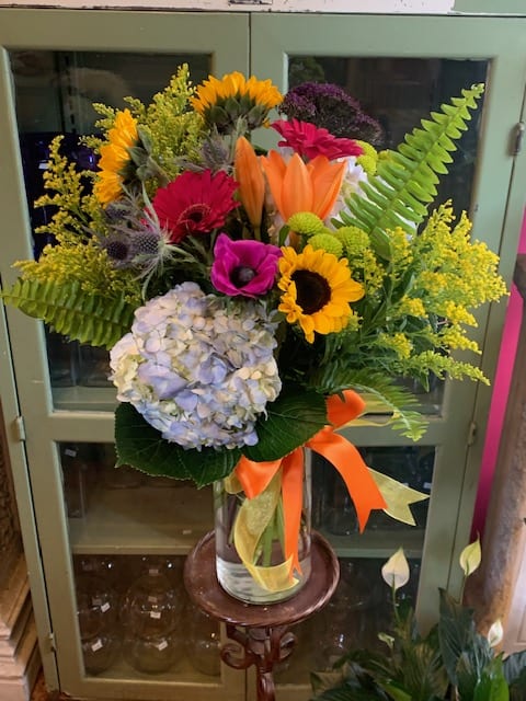 Tall mixed arrangement of sunflowers, lilies, gerbera daisies, hydrangea in a glass vase with orange ribbon