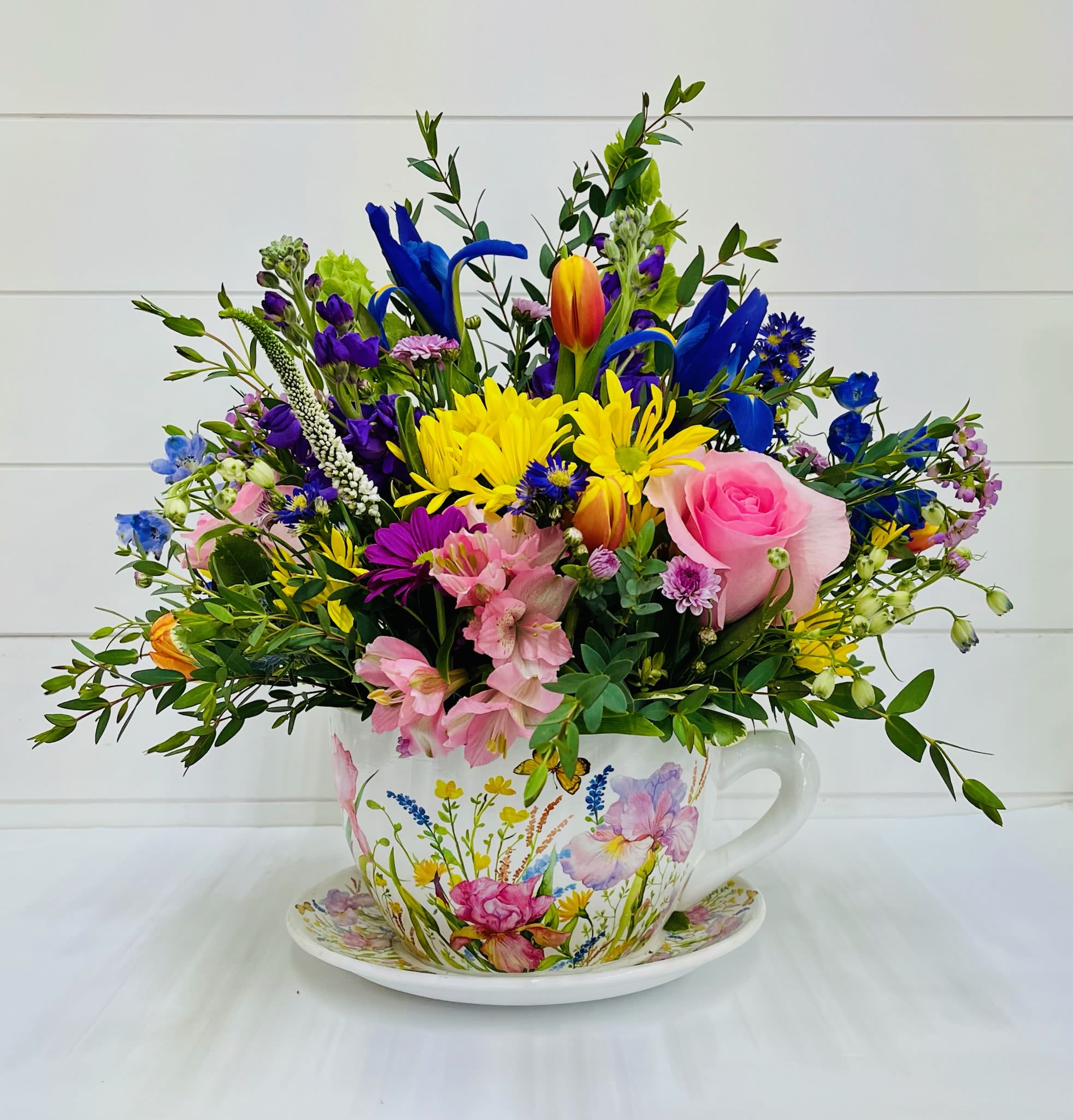 Colorful mixed flower arrangement in a floral teacup and saucer with pink, yellow, blue and purple blooms