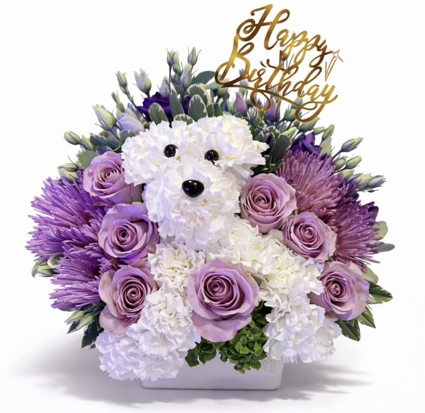 Birthday flower arrangement with a white carnation dog, lavender roses, and purple mums in a white container.