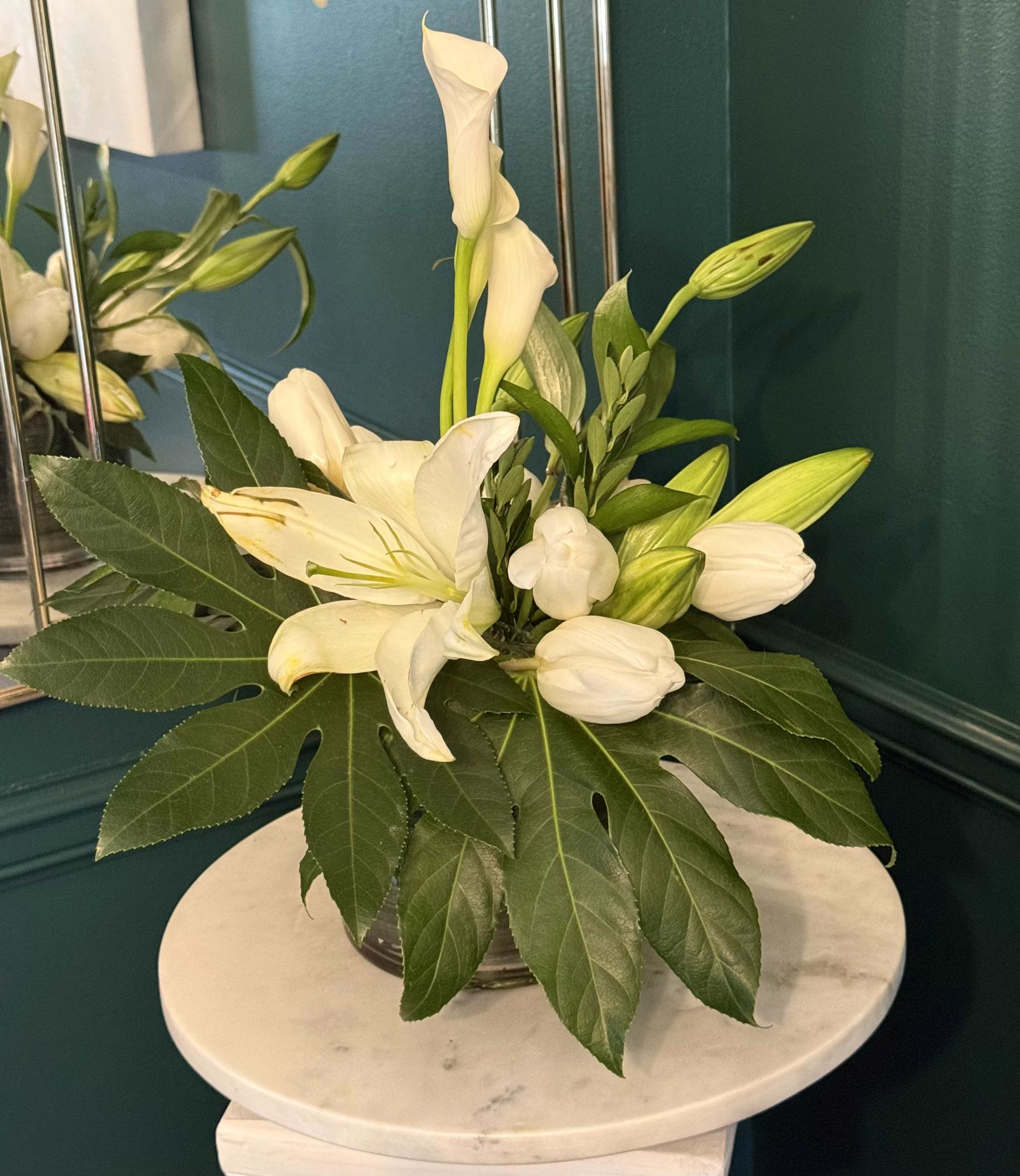 EMERALD ELEGANCE - A spare, modern silhouette of white calla lilies and tall Bells of Ireland makes this flower arrangement a calm, elegant choice for a Get Well gift or a simple "Thinking of You" moment. The vertical lines of the green Bells of Ireland lift the eye while the trumpet-shaped white callas offer a quiet, refined focal point-perfect for Just Because surprises, Thank You gestures, housewarming displays, or moments of sympathy.  The stems are gathered in a small, round clear glass vase so the straight stems are visible, reinforcing the arrangement's clean geometry. A white ribbon tied at the vase neck adds a soft finishing touch without distracting from the minimalist design. The green-and-white palette creates a serene presence that fits bedside tables, entry consoles, or a memorial setting.  Designed to make the recipient feel seen and comforted, this compact arrangement works anywhere a restrained, thoughtful floral statement is needed. Vase included; place in fresh water and trim stems if needed to extend bloom life.