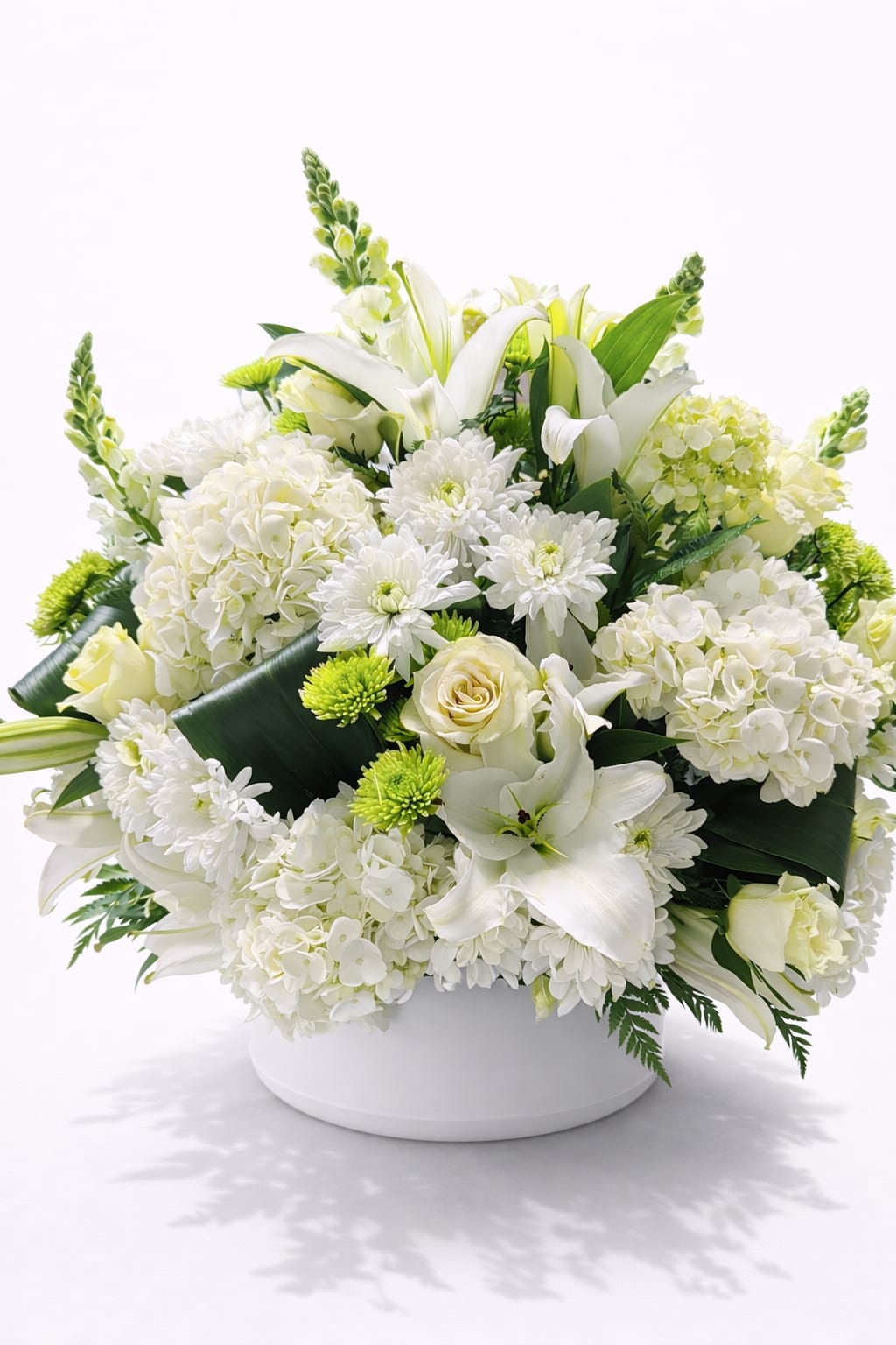 Low round arrangement of white lilies, roses, hydrangeas and green mums in a white bowl.