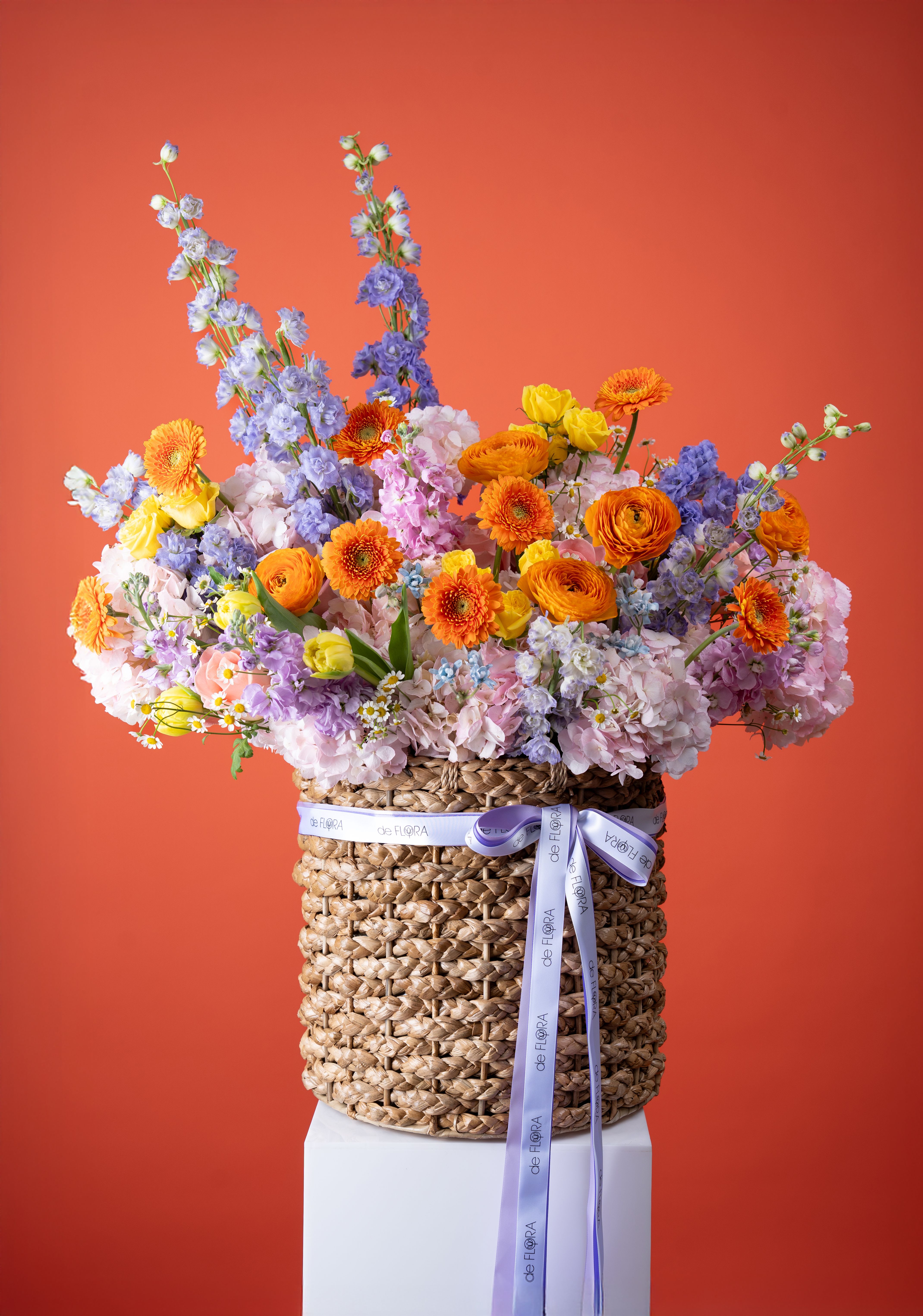 Sunset Garden Symphony - Bright orange gerbera daisies and ranunculus set the tone in this Sunset Garden Symphony flower arrangement, a mixed-color basket perfect as a birthday bouquet, Mother's Day surprise, or an uplifting "thinking of you" gift. Tall blue and lavender delphinium spires lift the composition, while soft pink and pastel hydrangeas and clusters of tiny white daisies and stock soften the palette-making it equally fitting for anniversaries, get well wishes, graduations, or just-because celebrations.  Arranged in a large, textured woven basket with a printed pale satin ribbon trailing down the side, the design balances bold warm tones of orange and yellow with cool lavenders, blues, and pinks for a loose, garden-inspired silhouette. Expect gerbera daisies, ranunculus, roses and spray roses, hydrangeas, delphinium, daisies and stock assembled with abundant, layered texture; the tall basket gives the piece vertical presence ideal for entryways or tabletops.  This lively, versatile arrangement makes someone feel noticed and celebrated-whether for Housewarming, Congratulations, Sympathy and Funeral, Thanksgiving, Christmas, or a seasonal Spring or Summer moment. It arrives ready to display in its woven container; for longest enjoyment, keep flowers in a cool spot away from direct heat and refresh the water as needed.