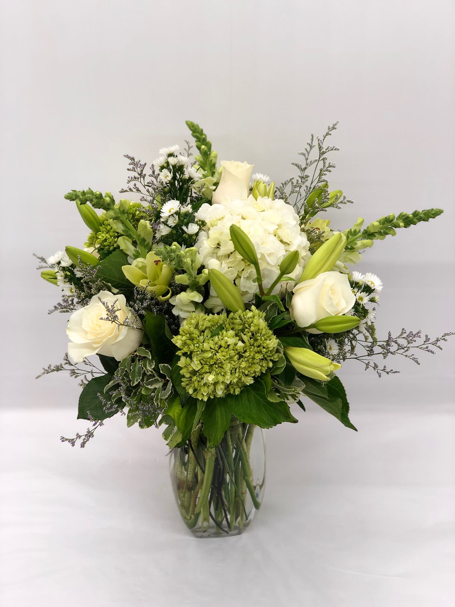 Tall arrangement of white and green roses, lilies, hydrangeas, and snapdragons in a clear glass vase.