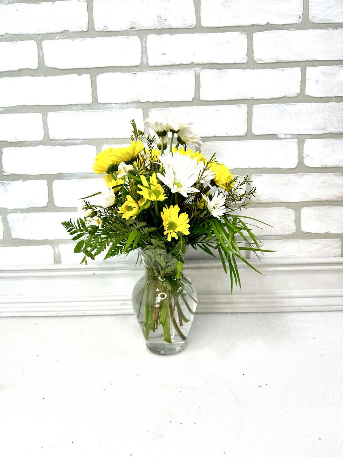 Small bouquet of yellow and white daisies arranged with greenery in a clear glass vase