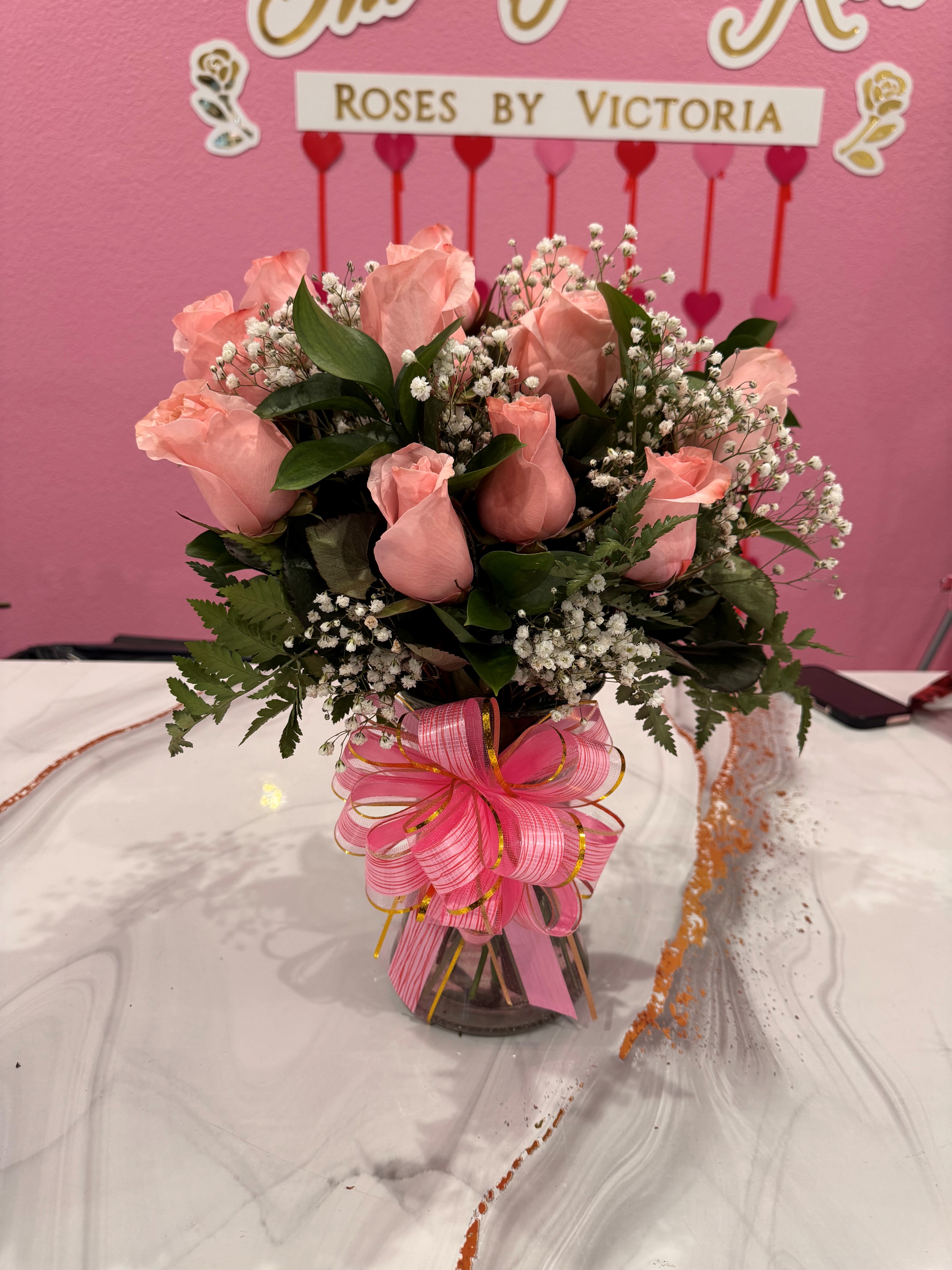 12ct Rose Vase Arrangement - Soft pink roses gathered in a compact, upright display create a sweet, romantic impression that makes the recipient feel genuinely celebrated - perfect as a housewarming gift or a newborn welcome. This 12-stem rose vase arrangement also reads beautifully for prom bouquets, a thank-you surprise, or a summer centerpiece when you want someone to feel cherished and seen.  Twelve soft pink roses are closely clustered with delicate white filler blooms for light contrast and airy texture, arranged in a clear glass vase for a classic, versatile look. A wide pink striped ribbon bow with subtle gold accents is tied at the vase neck to add a decorative, celebratory flourish. The arrangement's traditional styling and modest size work well as a tabletop centerpiece or an elegant bedside bouquet.  Send this to mark a new baby's arrival, a celebratory quinceañera, or to brighten a friend's new home - she'll feel warm and appreciated the moment it arrives. For best enjoyment, keep the vase topped up with fresh water and trim stems every few days to extend bloom life.