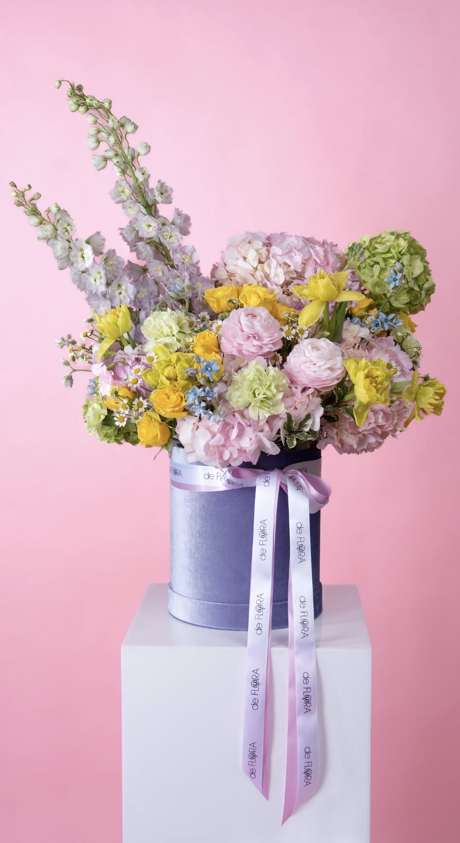 Blooming Elegance - A soft and uplifting composition designed to feel like a blooming spring garden. This elegant box features a harmonious blend of delicate pink ranunculus and hydrangea, sunny yellow spray roses and daffodils, airy delphinium, and charming chamomile accents. Hints of fresh green and touches of blue add depth and movement, while the modern deFLORA signature box with satin ribbon completes the look.  Light, joyful, and effortlessly chic — perfect for birthdays, celebrations, or simply to brighten someone’s day.