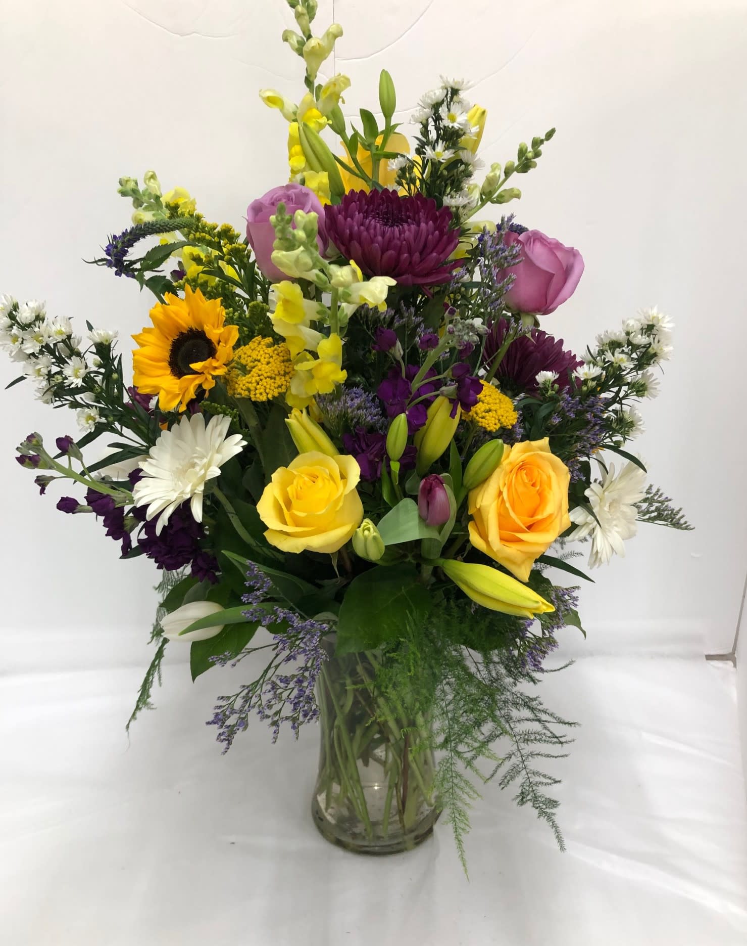 Tall mixed arrangement with yellow roses, sunflower, purple blooms and lilies in a clear glass vase