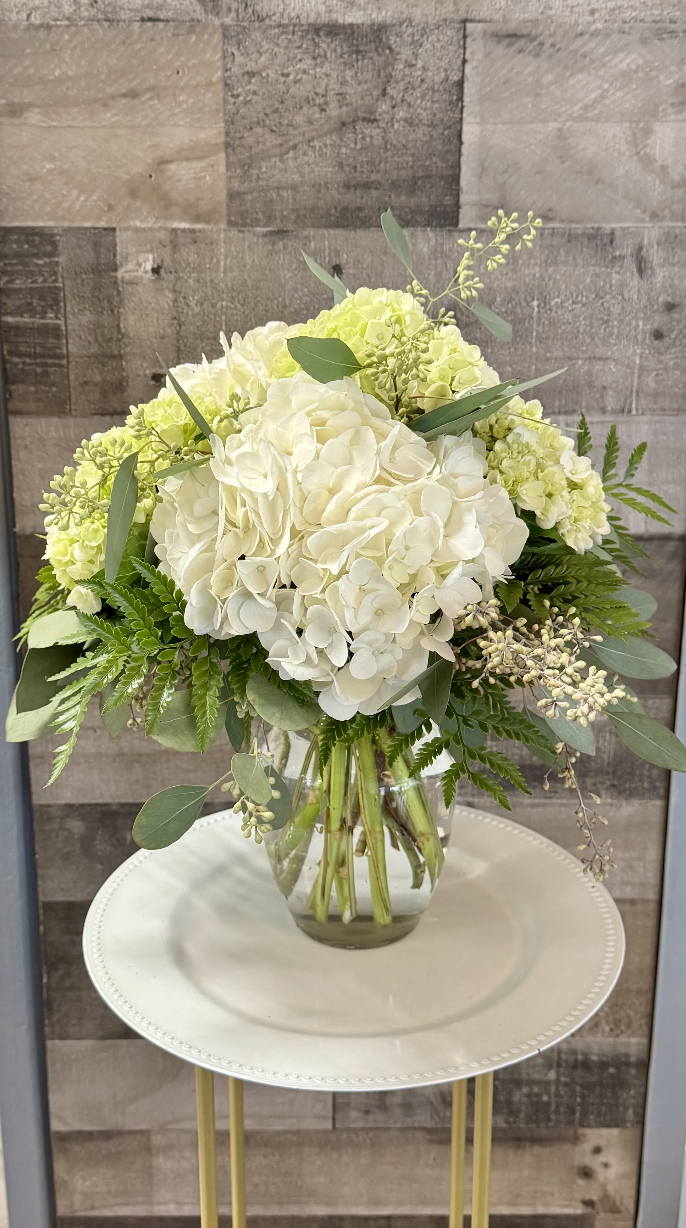 White and pale green hydrangea bouquet with lush foliage in a clear glass vase on a round white stand