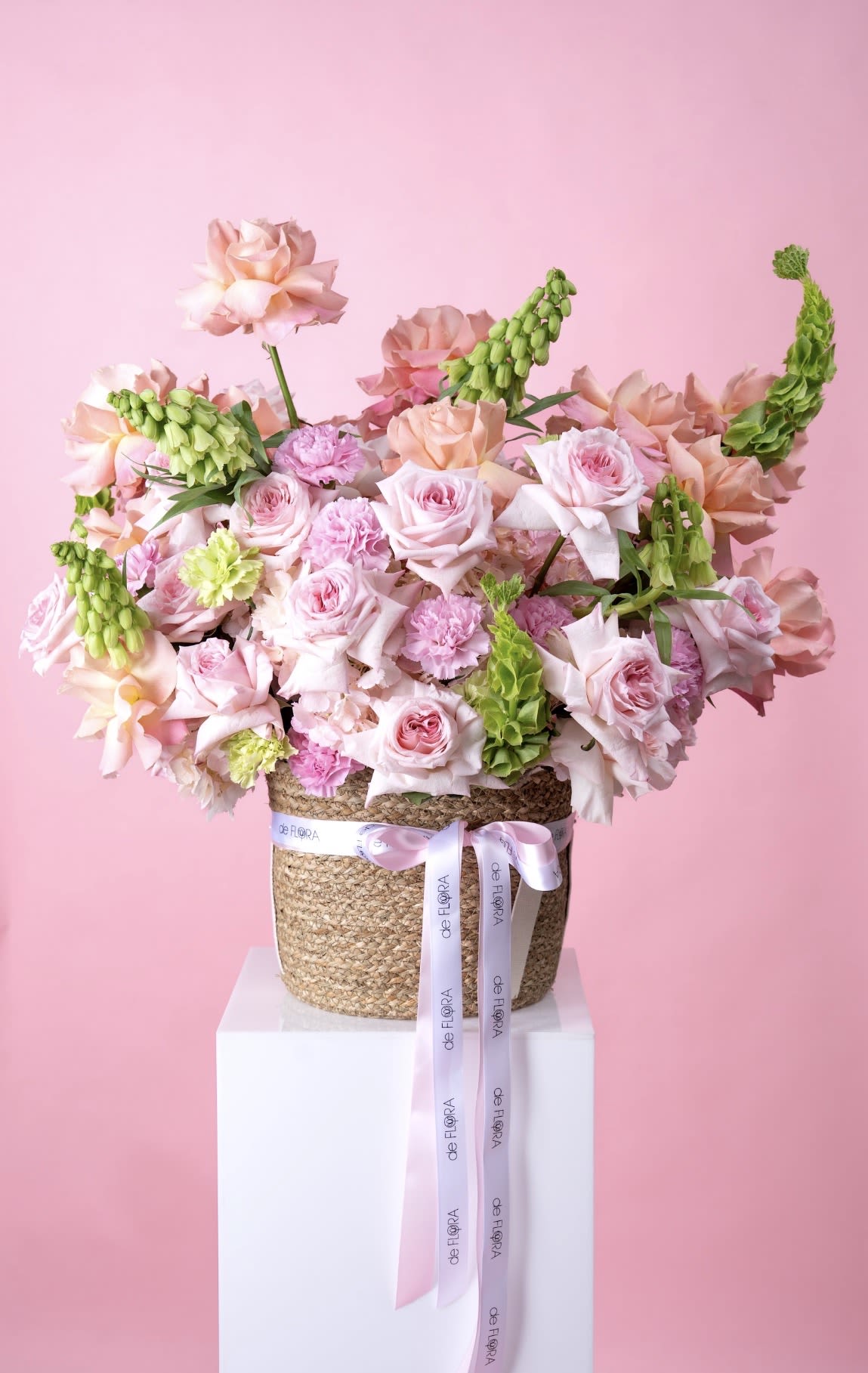 O’Hara Dream - Soft blush and peach roses mingle with ruffled pink carnations in a lush, full garden-style arrangement that feels like a gentle embrace - perfect as a birthday bouquet, anniversary flowers, or a Mother's Day gift. Tall, chartreuse Bells of Ireland rise above the blooms to add graceful height and a crisp green contrast that brightens the pastel palette; the overall effect is romantic, feminine, and quietly celebratory so the person you're thinking of will feel truly seen and cherished.  The blooms are densely gathered in a natural woven basket for a casual yet refined presentation, finished with long printed satin ribbons and a white branded ribbon that drape down the front for a polished touch. Colors are soft pink, peach, green and white; flowers include roses, carnations, Bells of Ireland and complementary seasonal stems for texture and movement. This arrangement arrives ready to display on a mantel or table - ideal for anniversaries, new baby celebrations, get well wishes, or anytime you want to send warmth and affection.