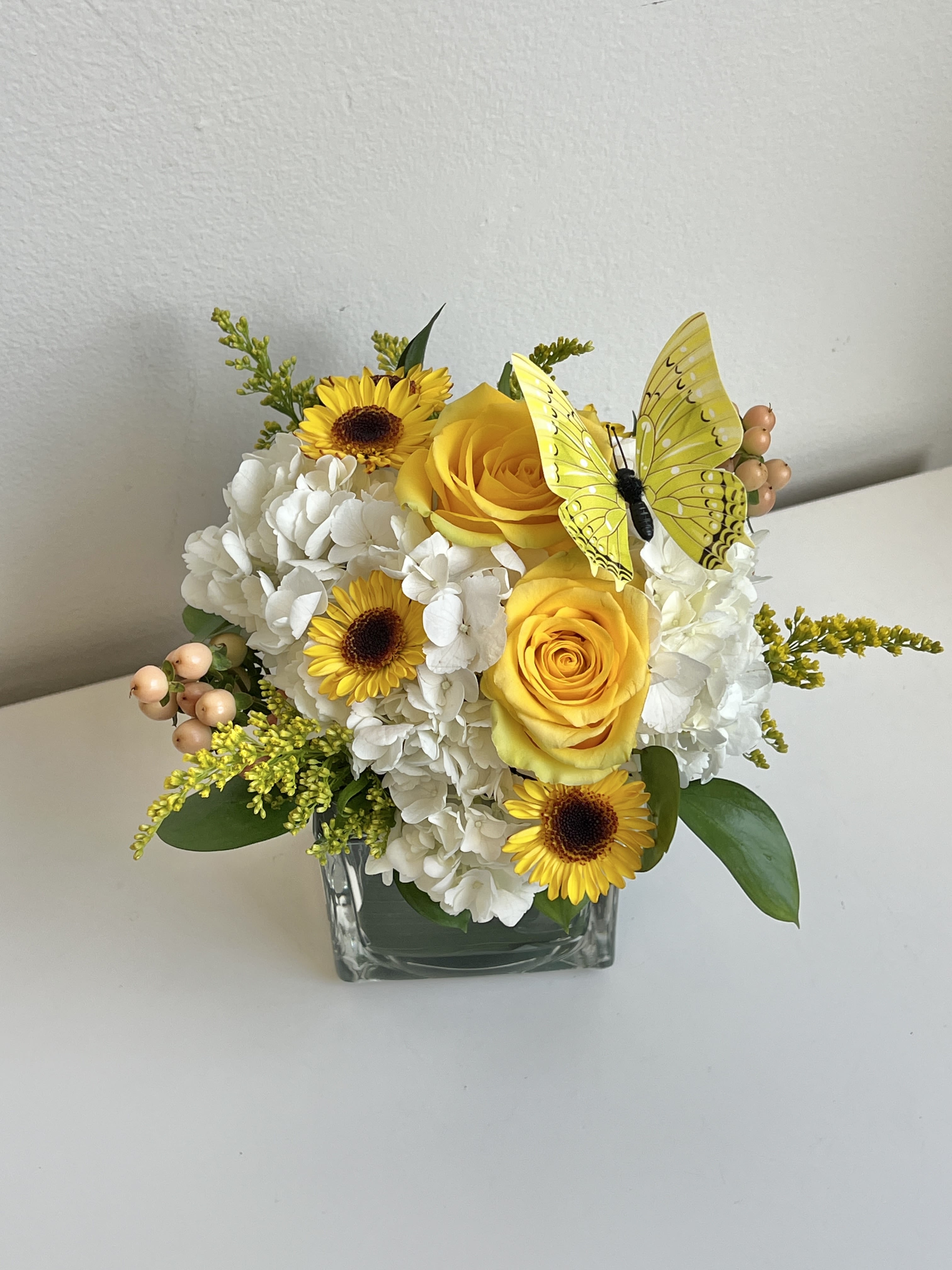 Compact yellow and white arrangement with roses, hydrangea, and daisies in a glass cube with a decorative butterfly