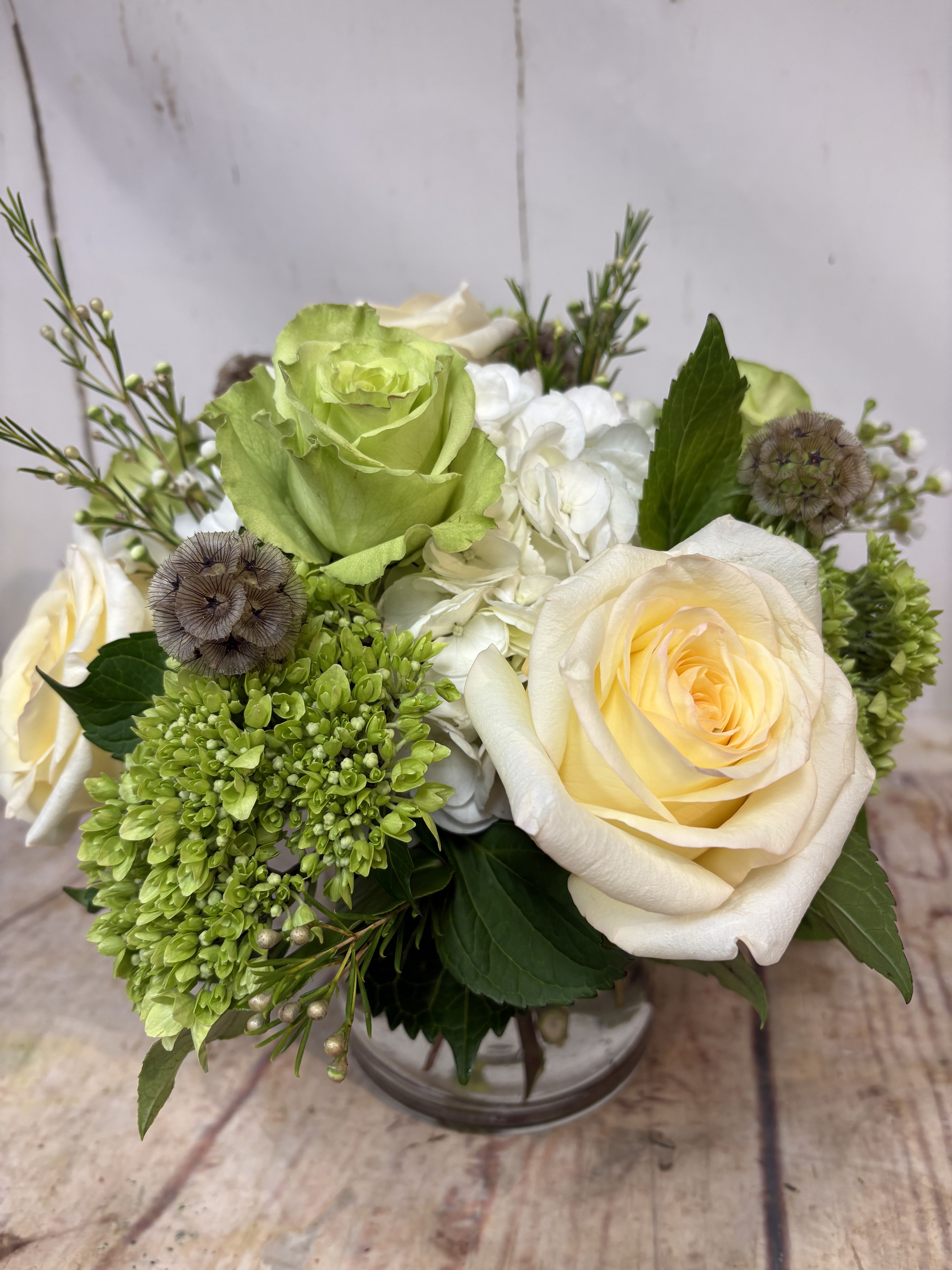 Low arrangement of cream roses, green blooms, and white hydrangeas in a clear glass vase
