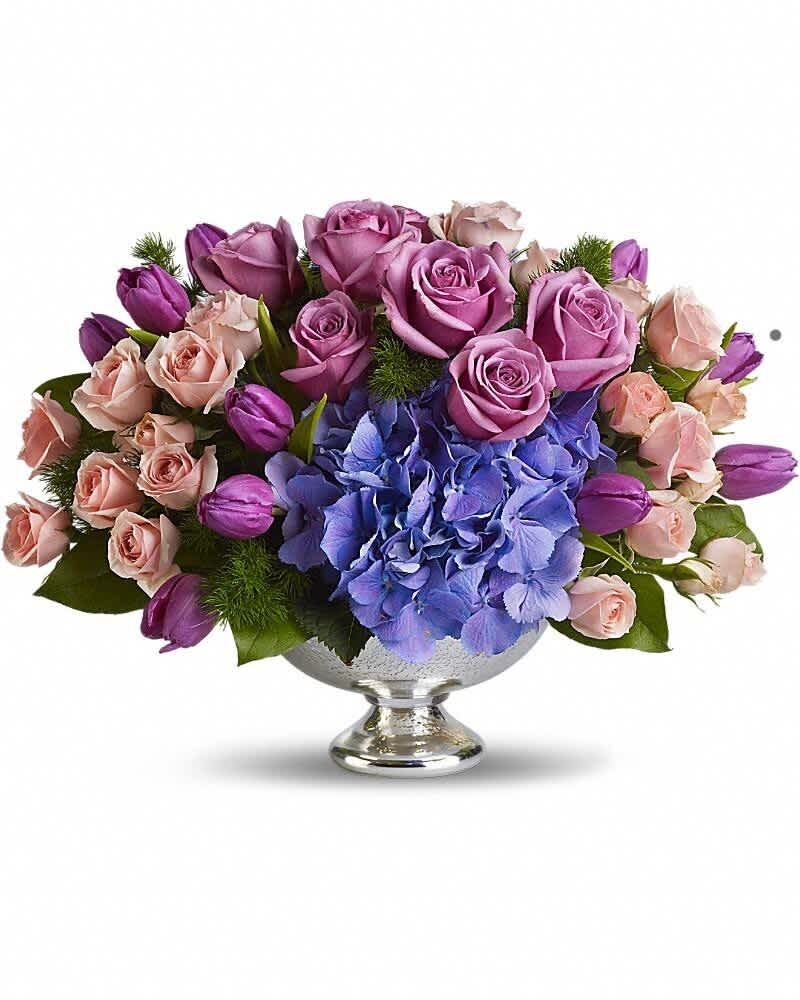 Low centerpiece of purple roses, pink spray roses, tulips, and blue hydrangeas in a silver pedestal bowl