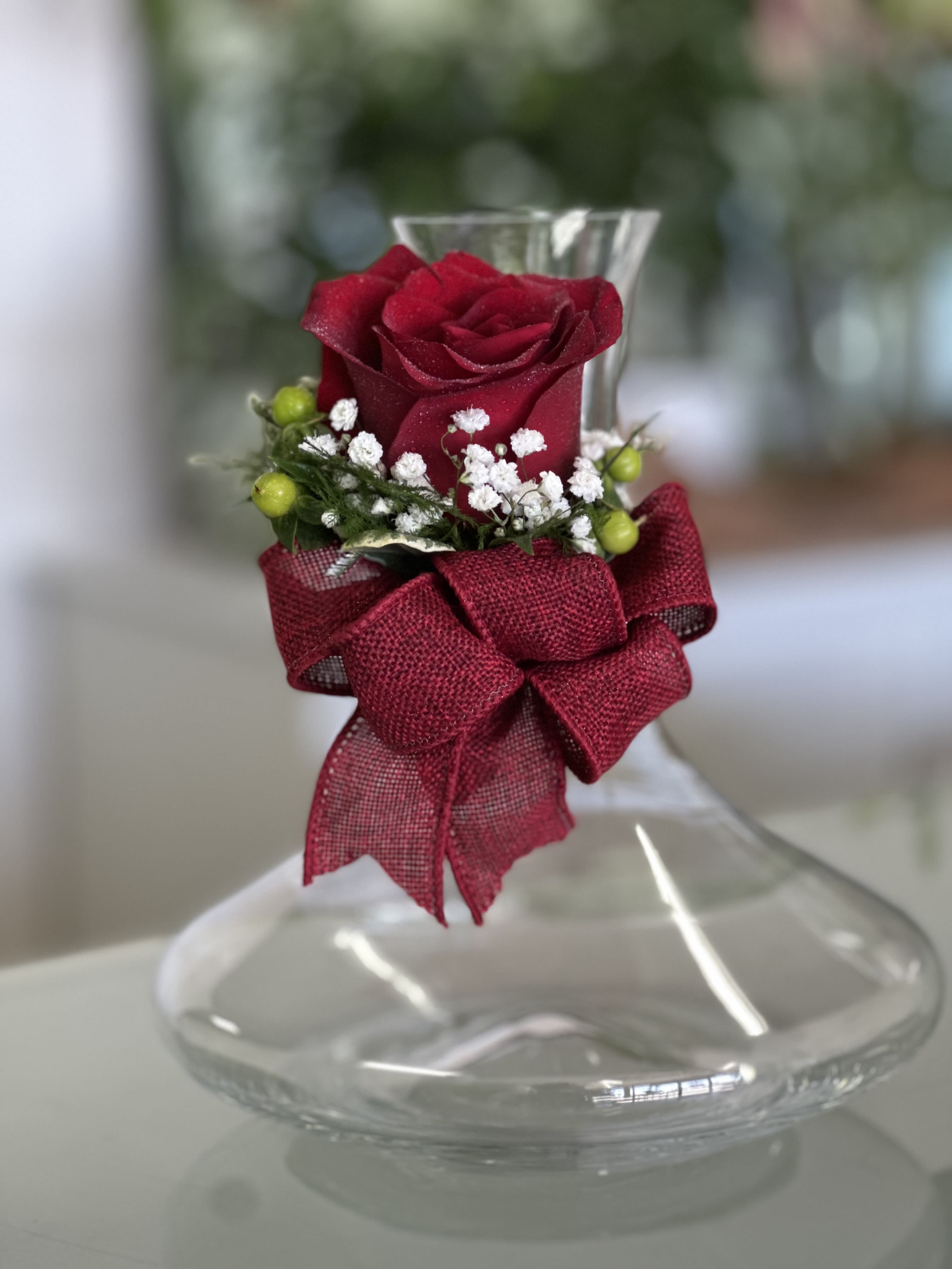Clear glass vase with a single red rose, white filler flowers, green berries, and a red bow at the neck.