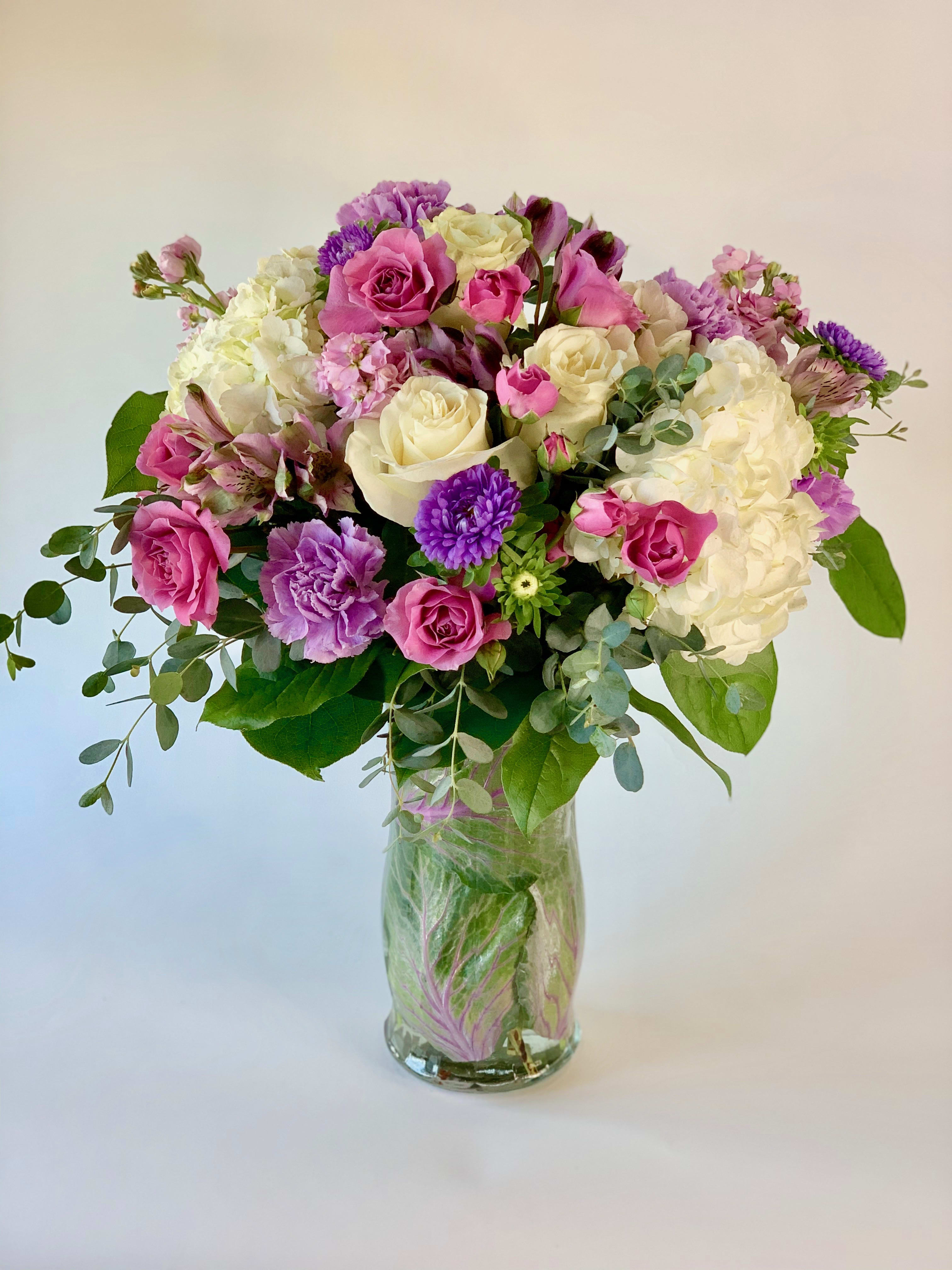 Mixed bouquet of pink, white, and purple flowers in a clear glass vase lined with green leaves