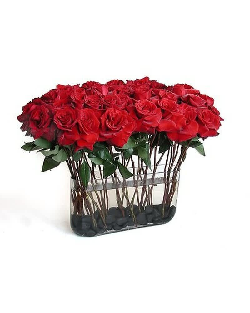 Rectangular vase filled with many red roses arranged in tight rows over black stones.