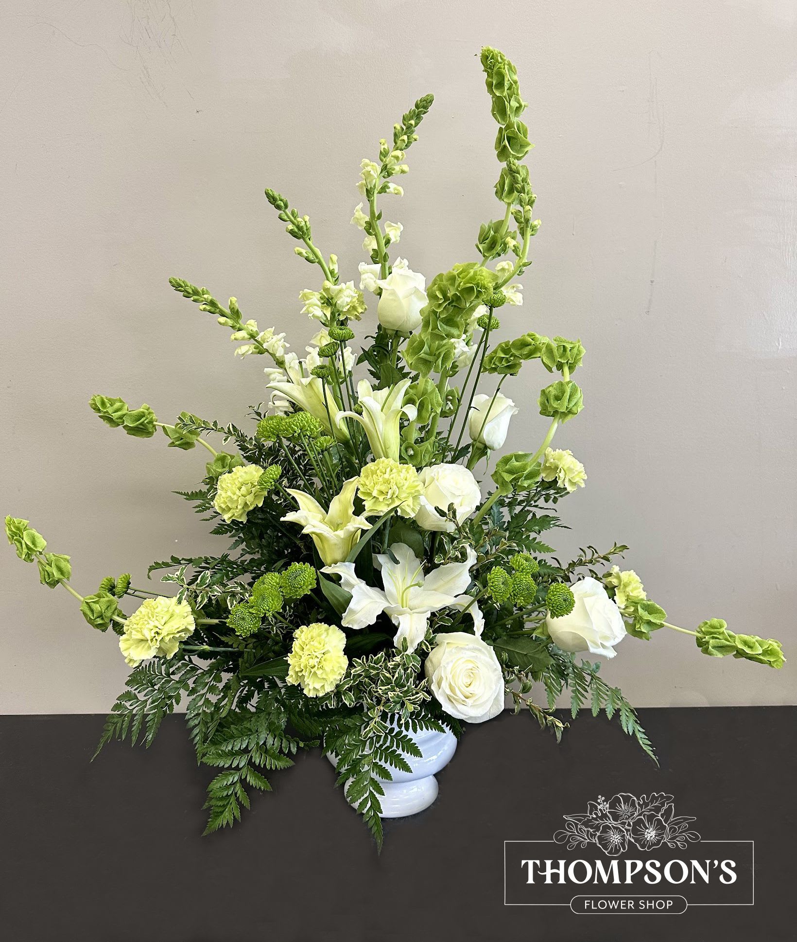 Tall white and green arrangement with lilies, roses, carnations, and Bells of Ireland in a white vase