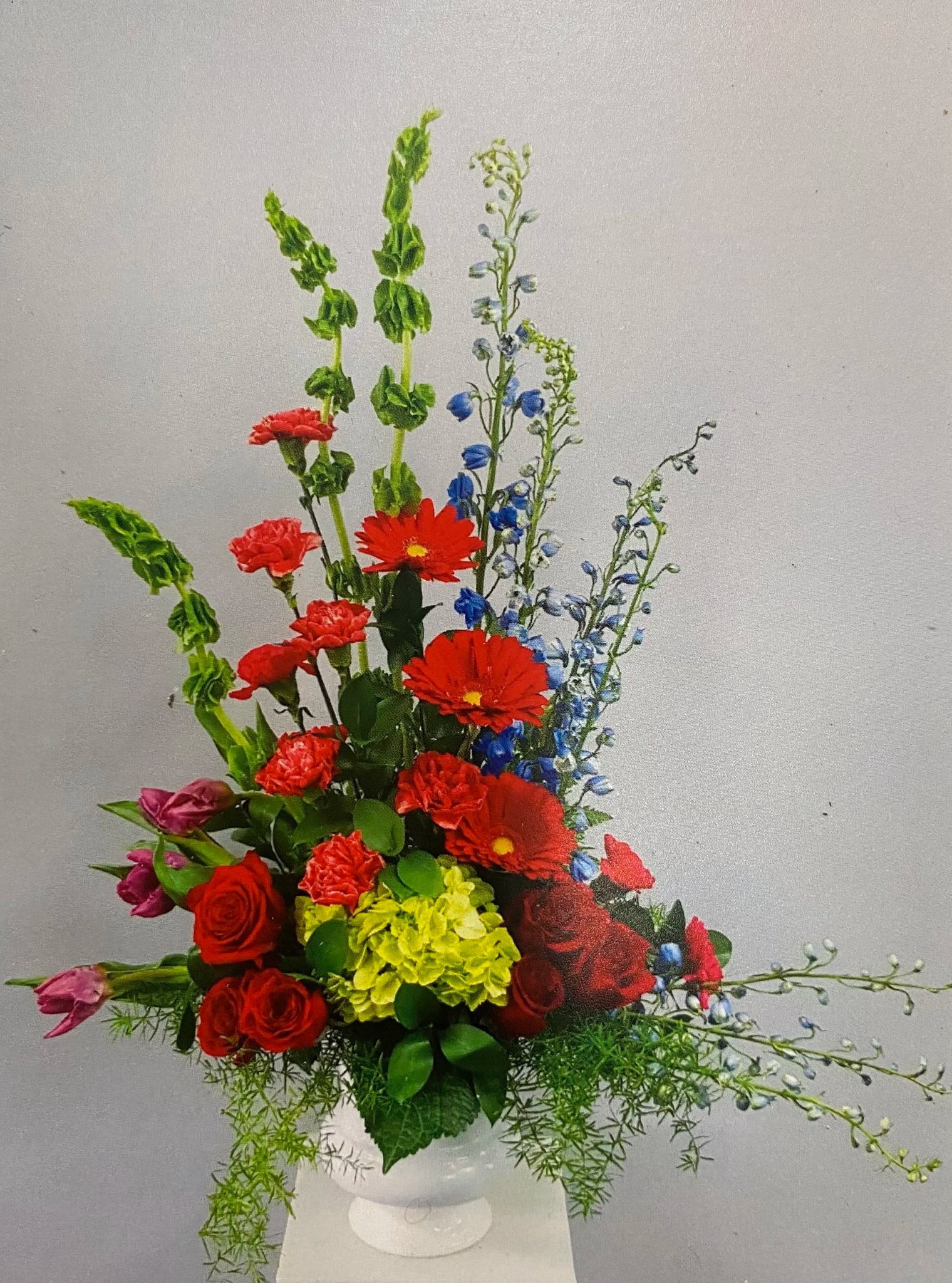 Tall mixed arrangement with red roses, gerbera, blue delphinium, and green hydrangea in a white vase