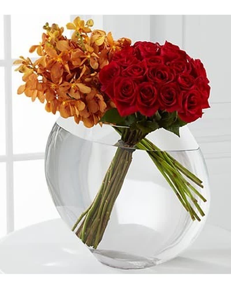 Red roses and orange orchids arranged at an angle in a round clear glass vase