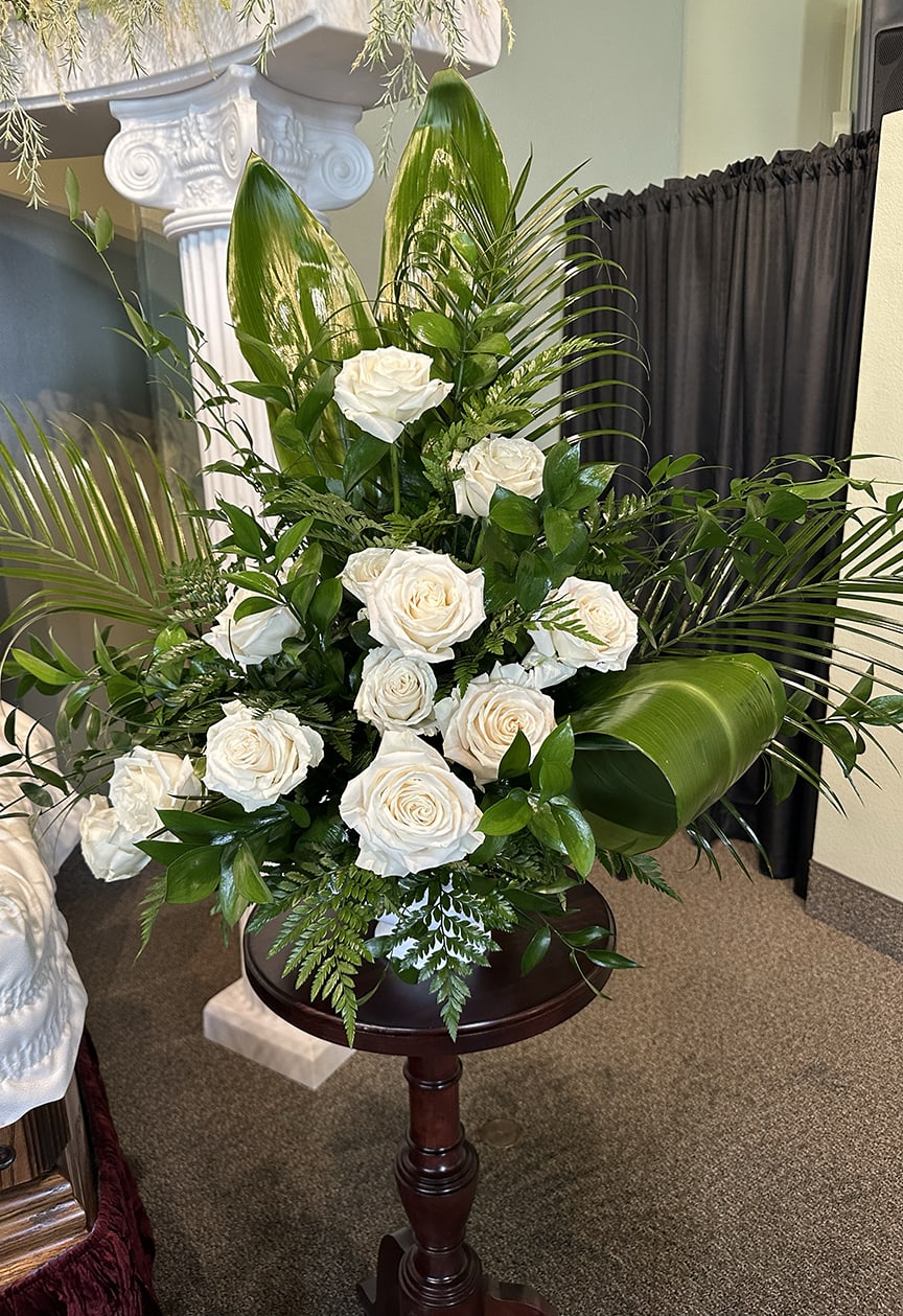 Tall arrangement of white roses with large green tropical leaves on a wooden pedestal.