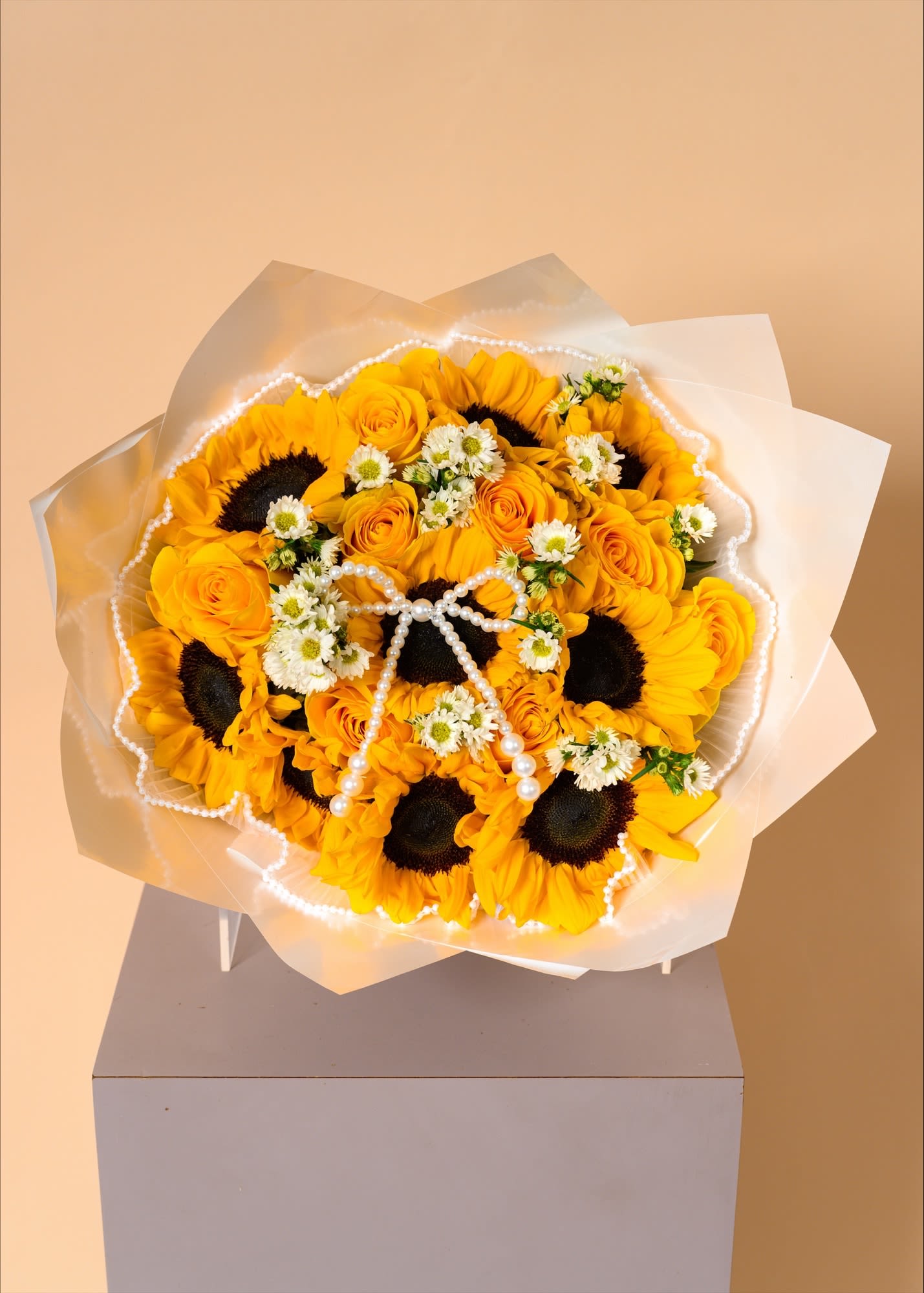 Golden Radiance Bouquet - A stunning bouquet of vibrant sunflowers and golden roses, delicately accented with petite white blooms for a soft, romantic contrast. Wrapped in elegant translucent paper and finished with a pearl detail, this bouquet exudes warmth, joy, and refined luxury—perfect for brightening any special moment.