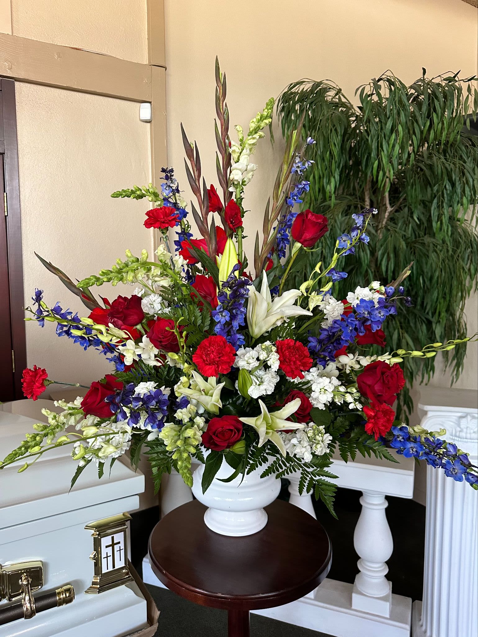 Tall red, white, and blue funeral arrangement with roses, lilies, carnations, and delphinium in a white urn.