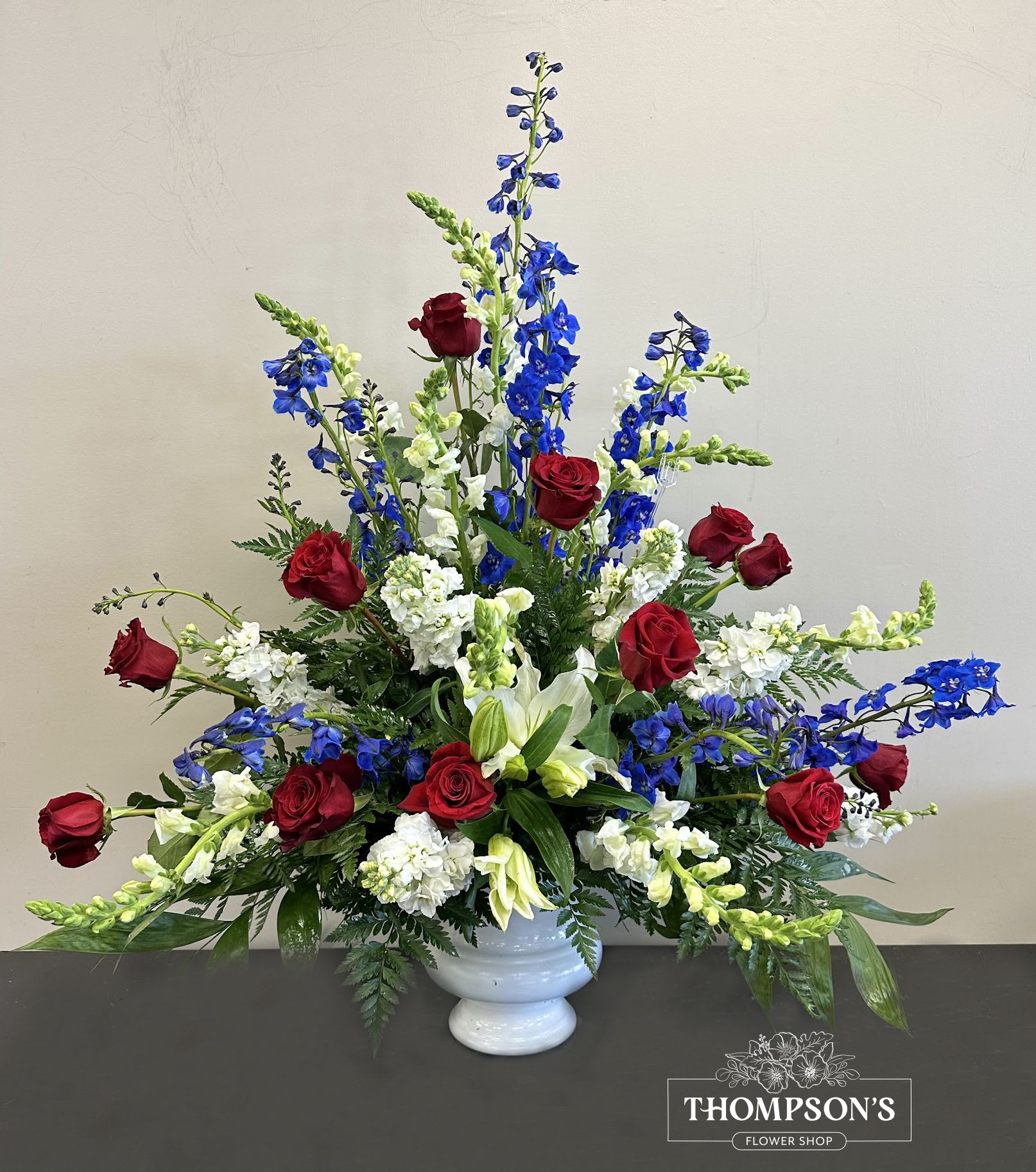 Tall red, white, and blue arrangement with roses, lilies, and delphinium in a white urn vase