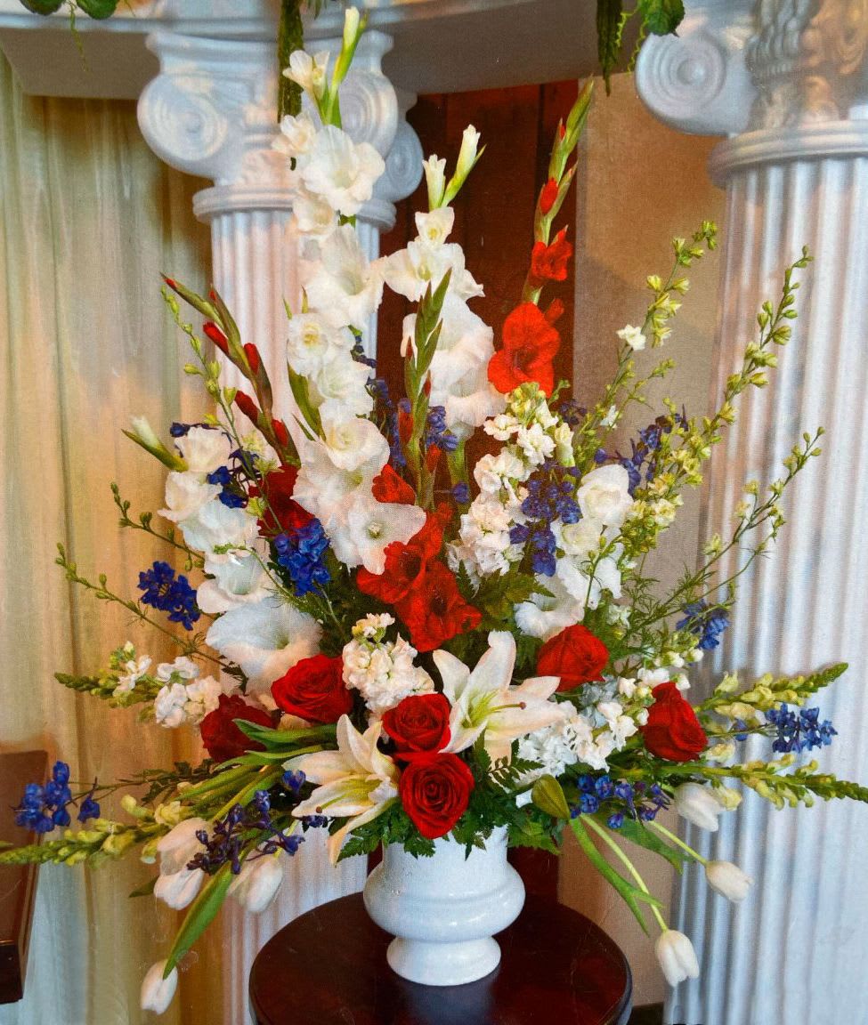Tall red, white, and blue floral arrangement with roses, lilies, and delphinium in a white urn vase