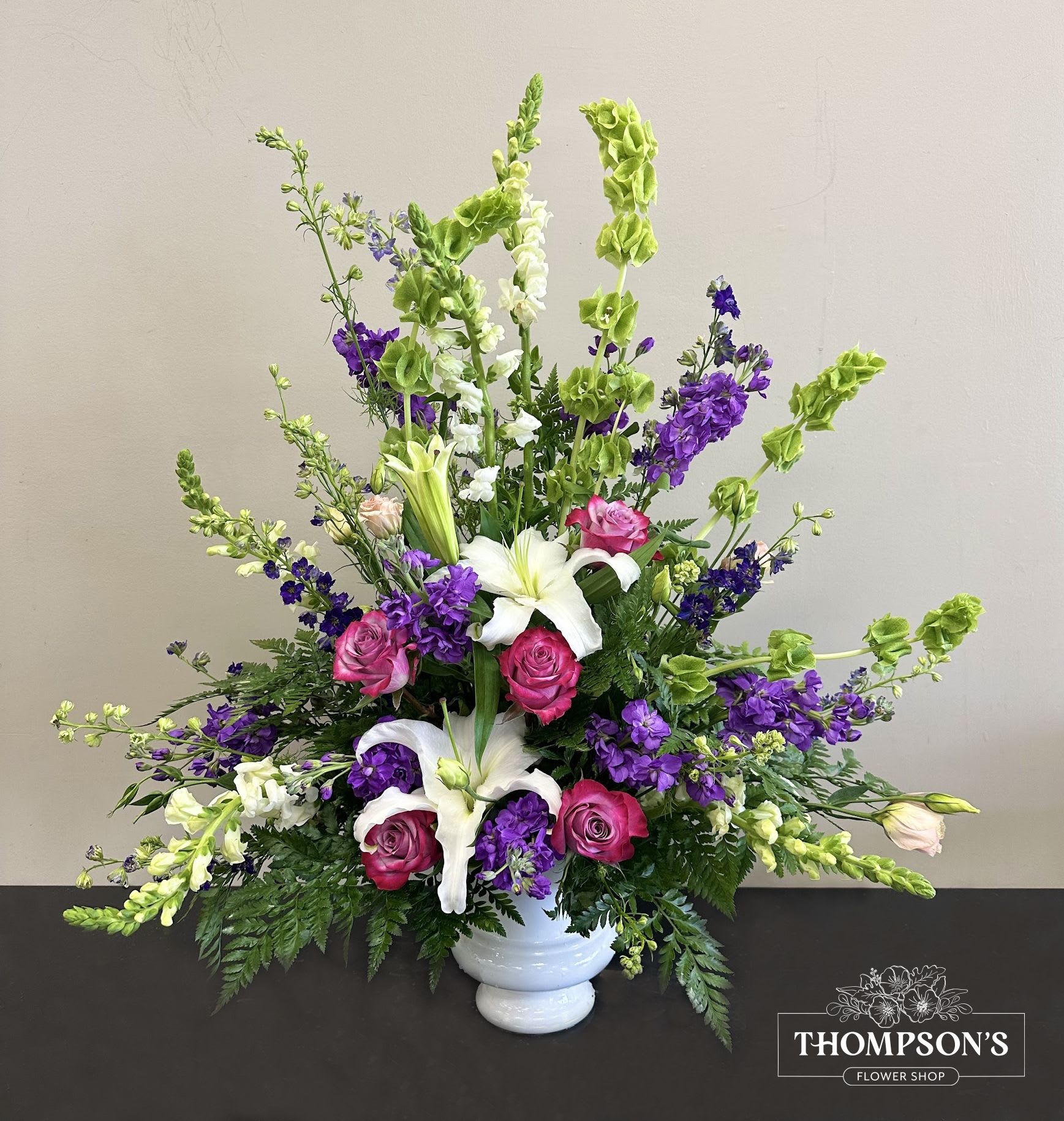 Tall fan-shaped arrangement with white lilies, pink roses, purple blooms, and green bells of Ireland in a white urn.