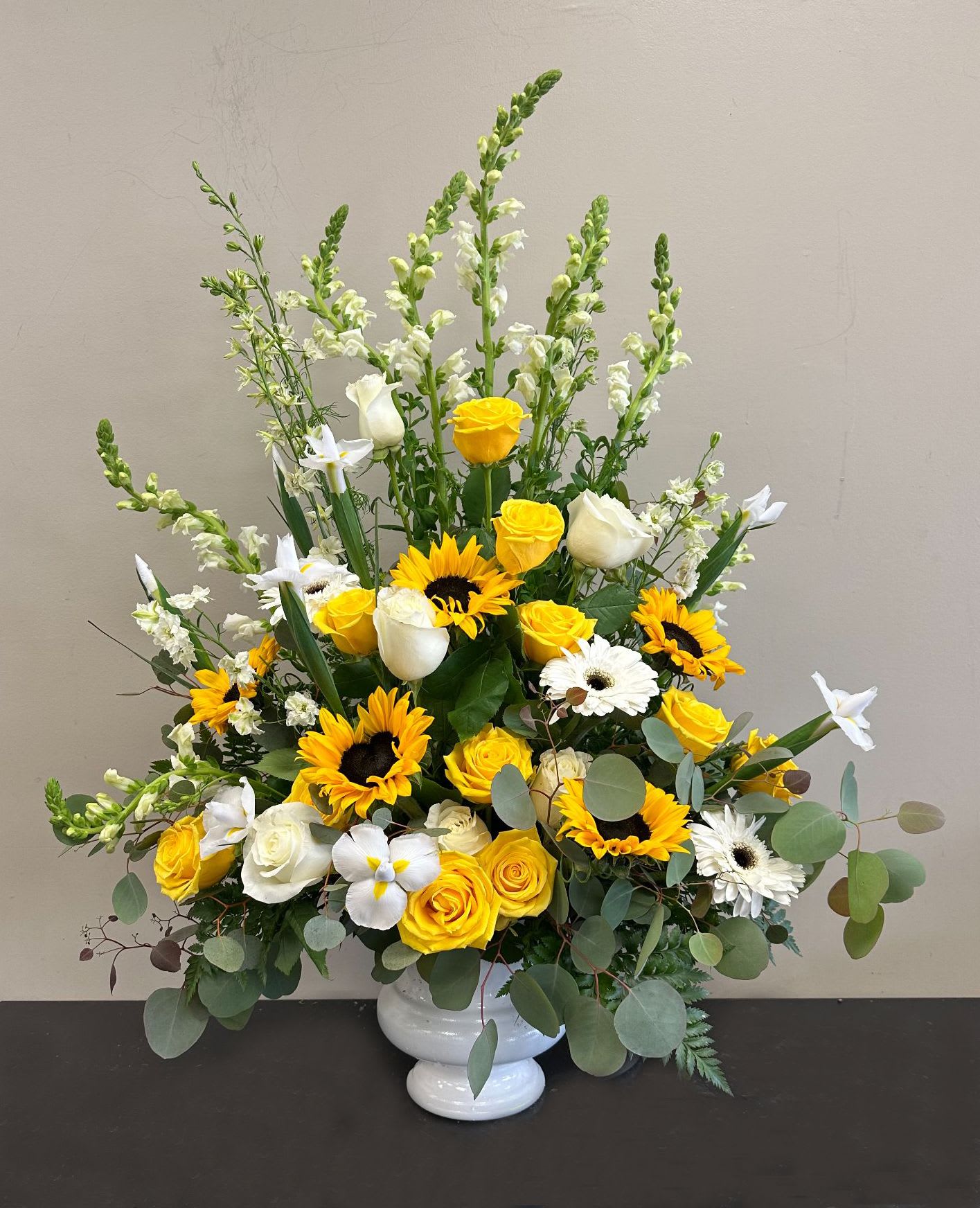 Tall yellow and white arrangement with sunflowers, roses, daisies and snapdragons in a white pedestal vase
