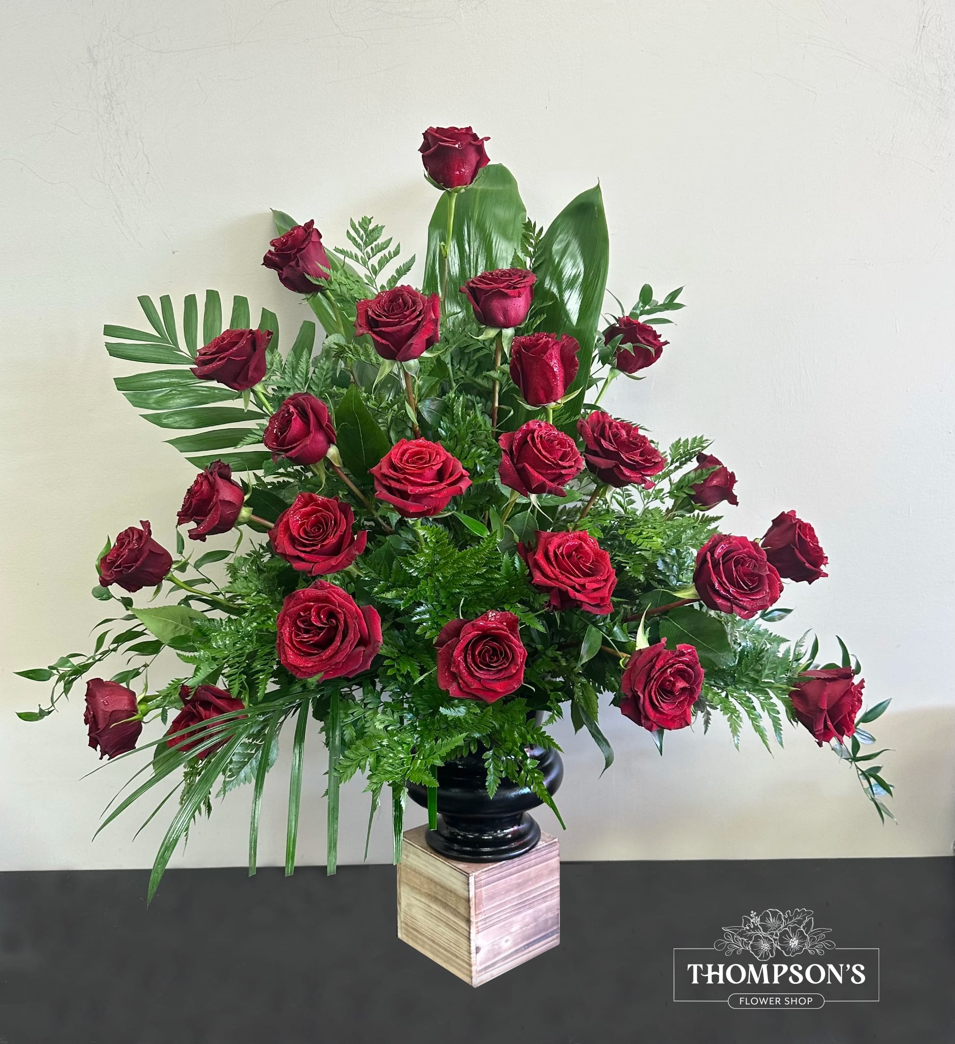 Tall arrangement of red roses with lush greenery in a black vase on a wooden pedestal.