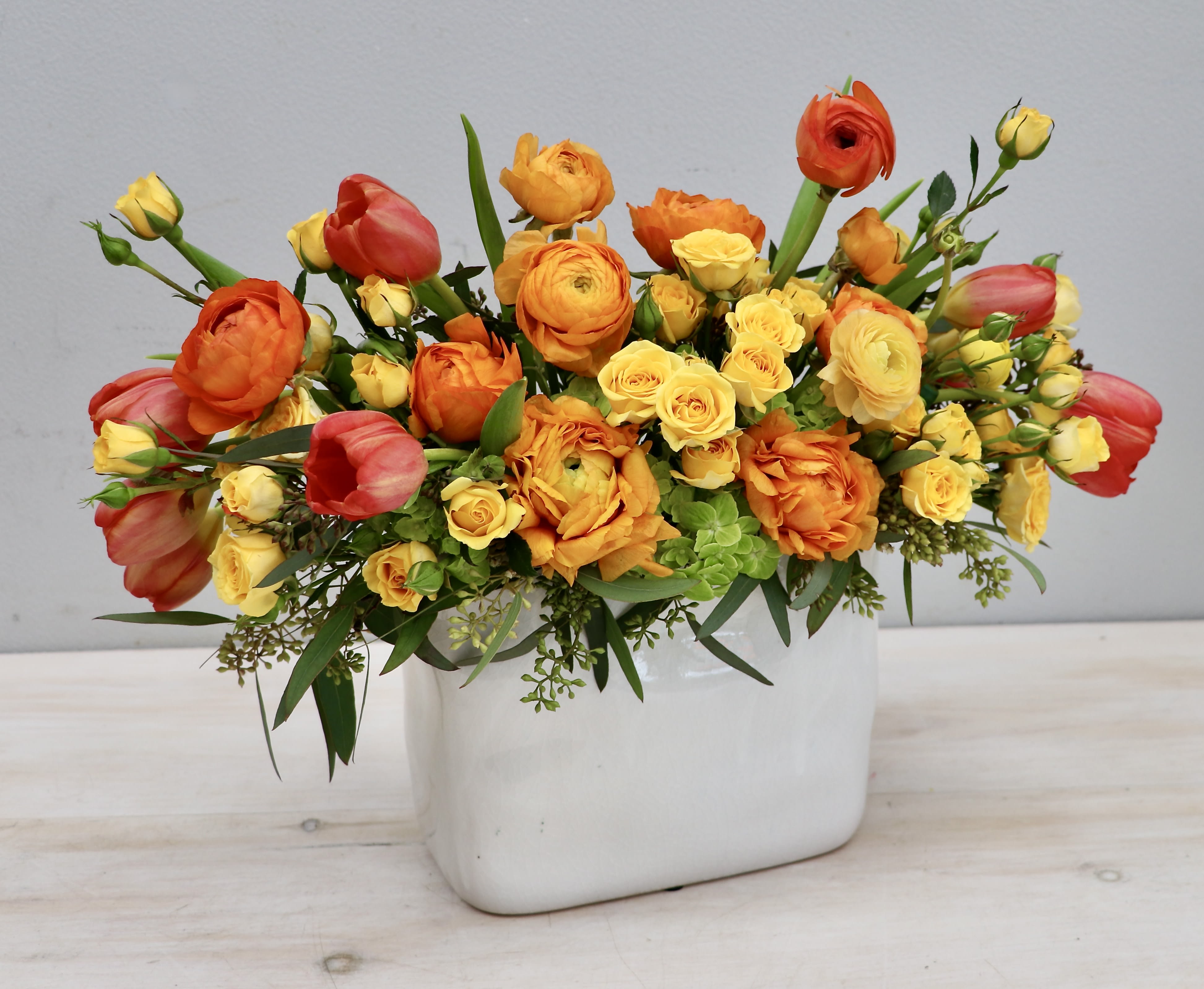Low arrangement of orange ranunculus, yellow spray roses, and coral tulips in a white rectangular vase
