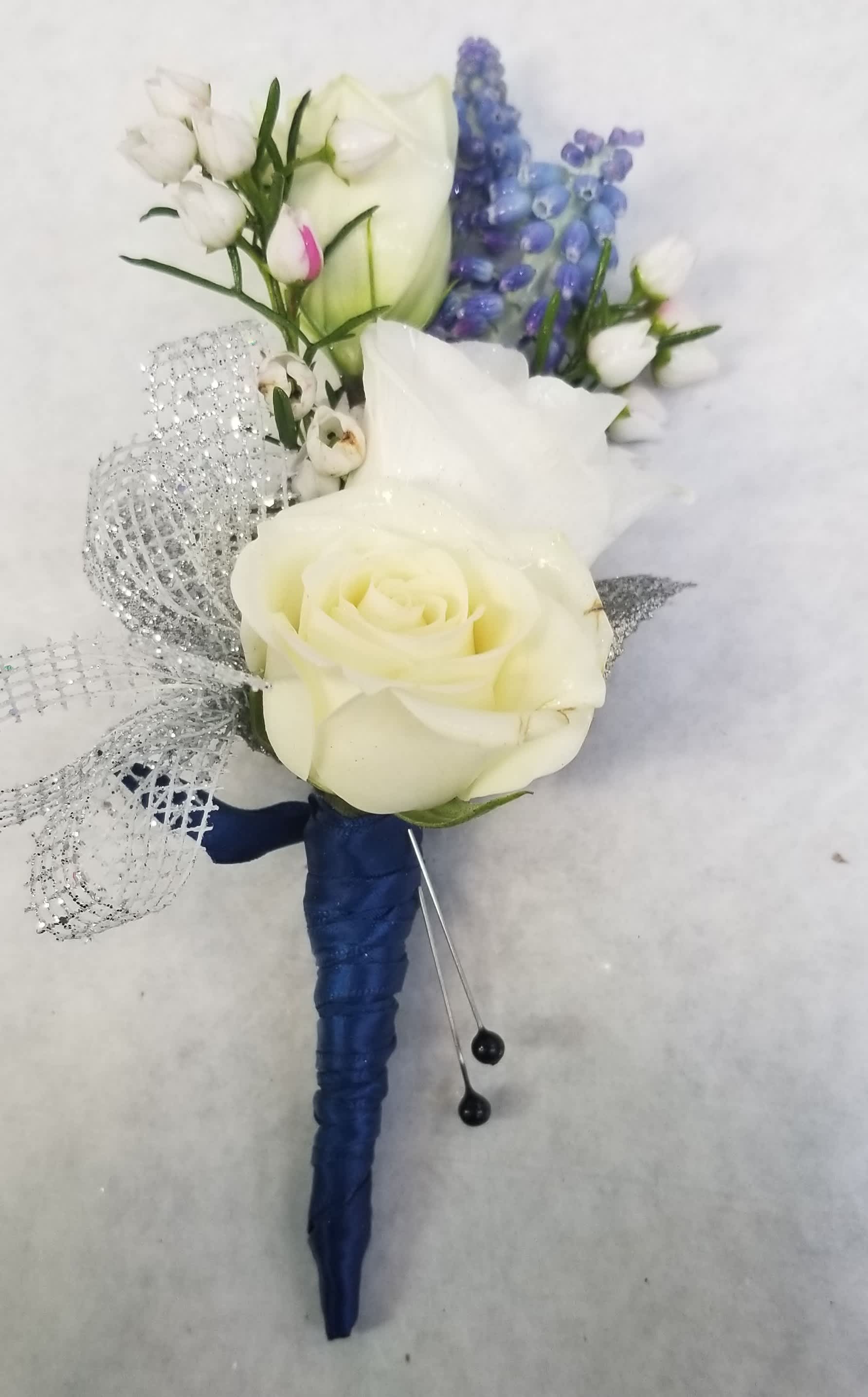 White rose boutonniere with blue ribbon wrap, silver bow, and small purple accent flowers.